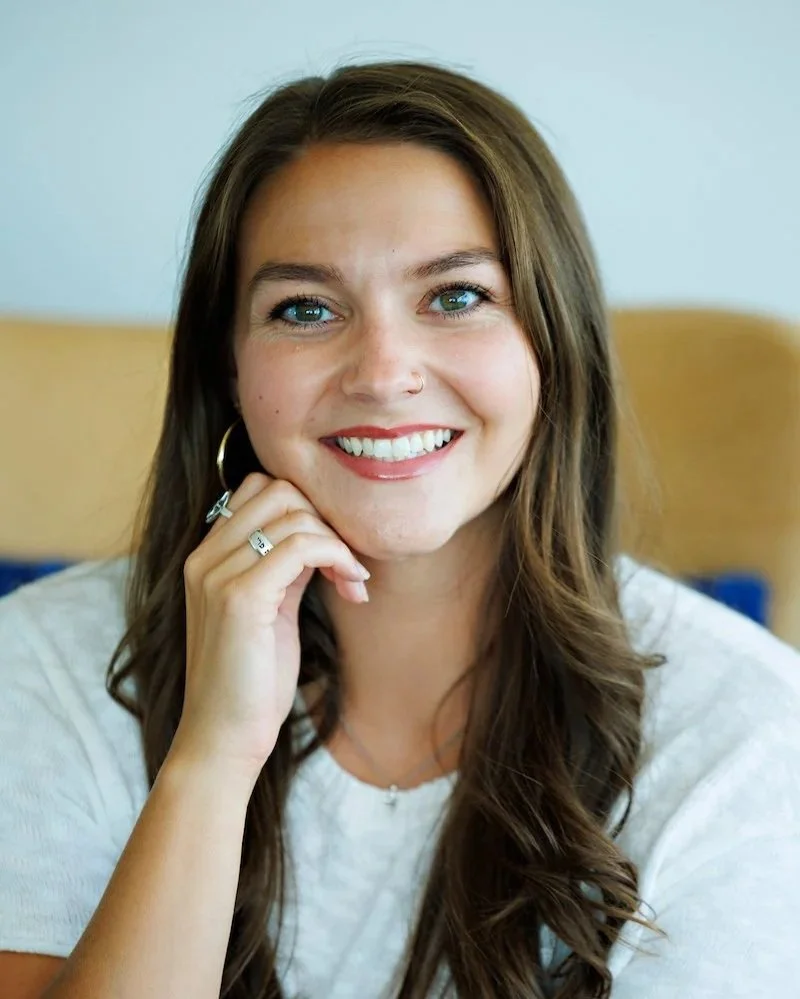 A woman with long brown, wet hair, smiling, wearing hoop earrings, a small nose ring, and a silver cross necklace, in a bright modern office with white shelves, plants, and a framed motivational poster.