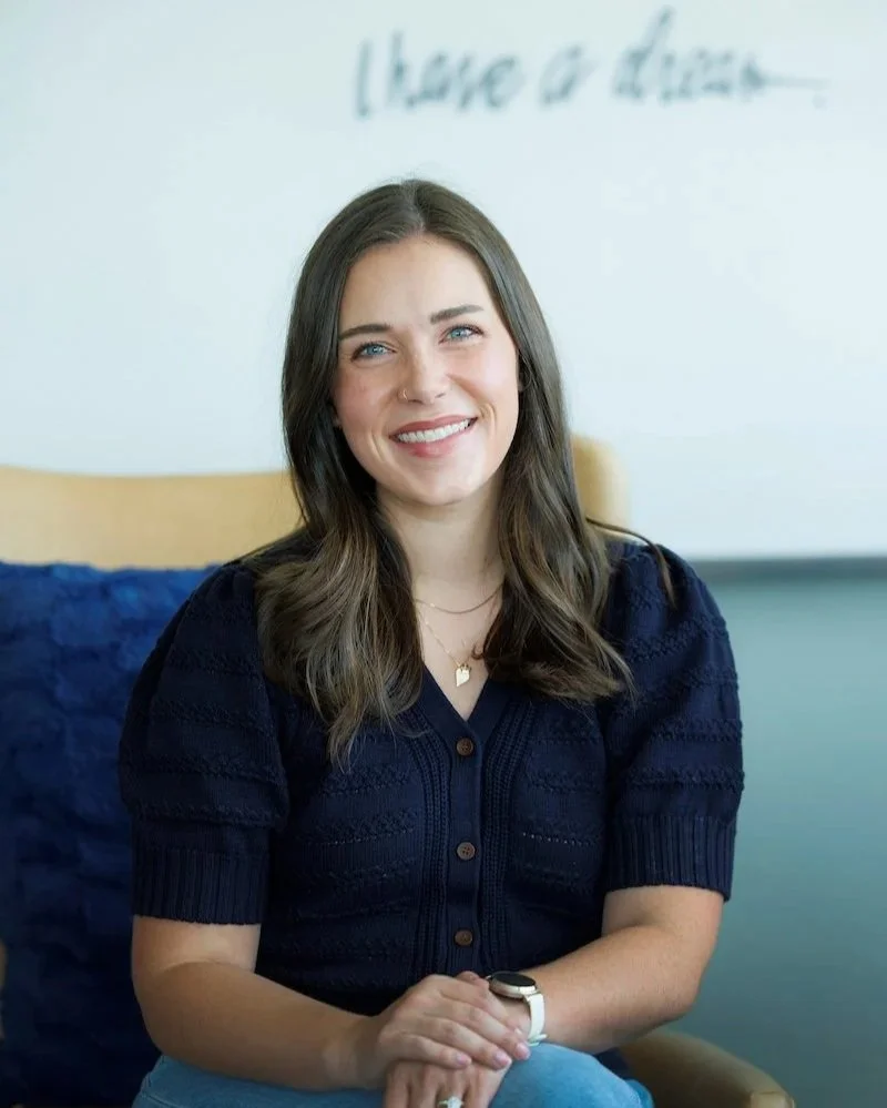 Close-up of a smiling woman with dark brown hair, blue eyes, wearing a white shirt with the top button undone, standing in front of a blurred bookshelf background.