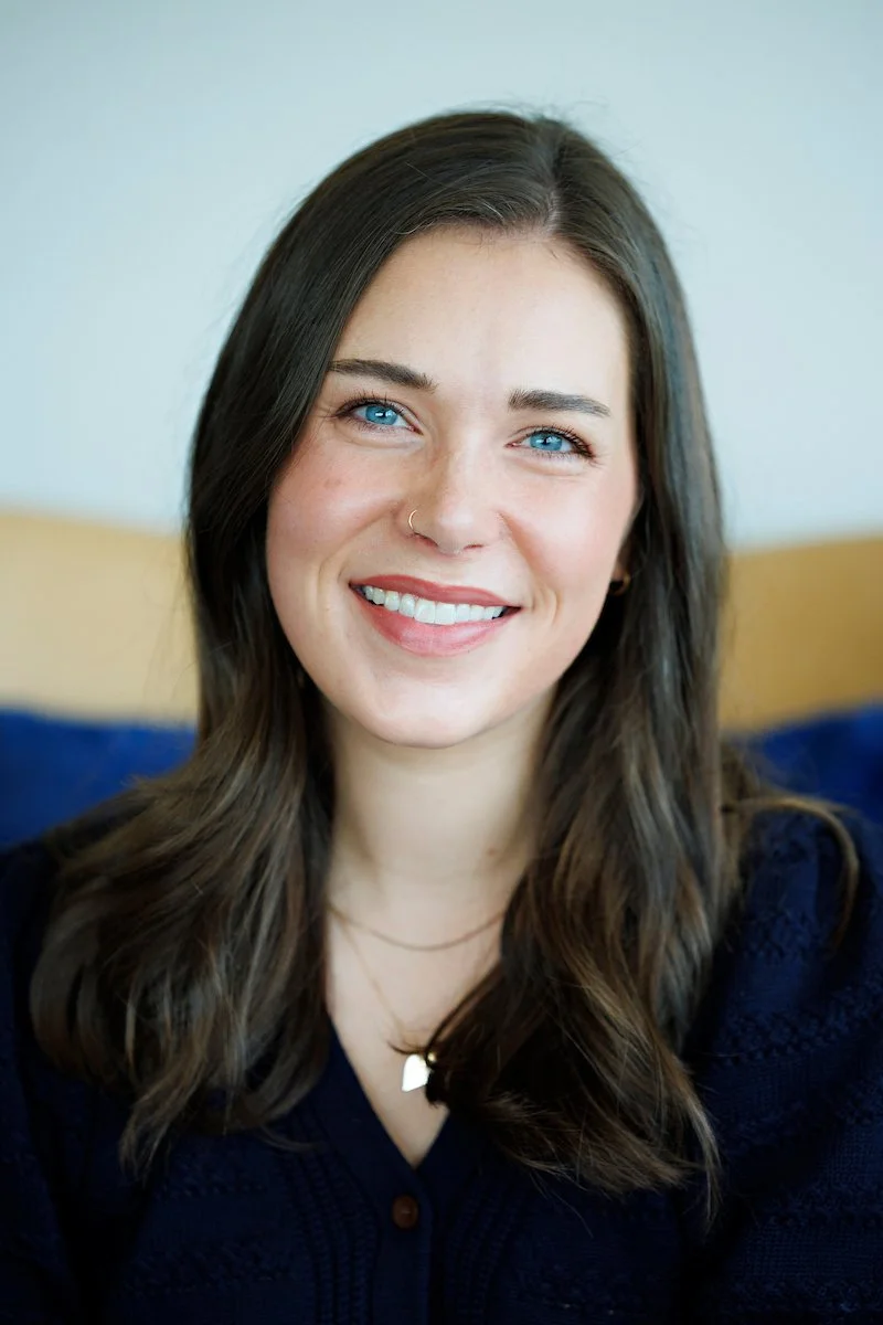A young woman with long brown hair, blue eyes, and a nose ring, smiling at the camera. She is wearing a dark navy sweater and a small necklace. The background features a light-colored wall and a blue cushion or blanket.