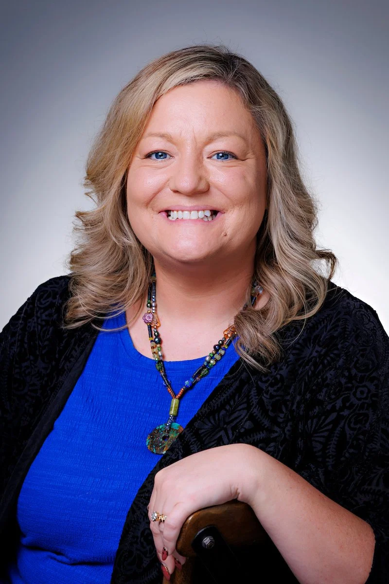 A smiling woman with blonde hair, wearing a blue top, black patterned jacket, colorful beaded necklace, a ring, and resting her arm on a wooden chair.