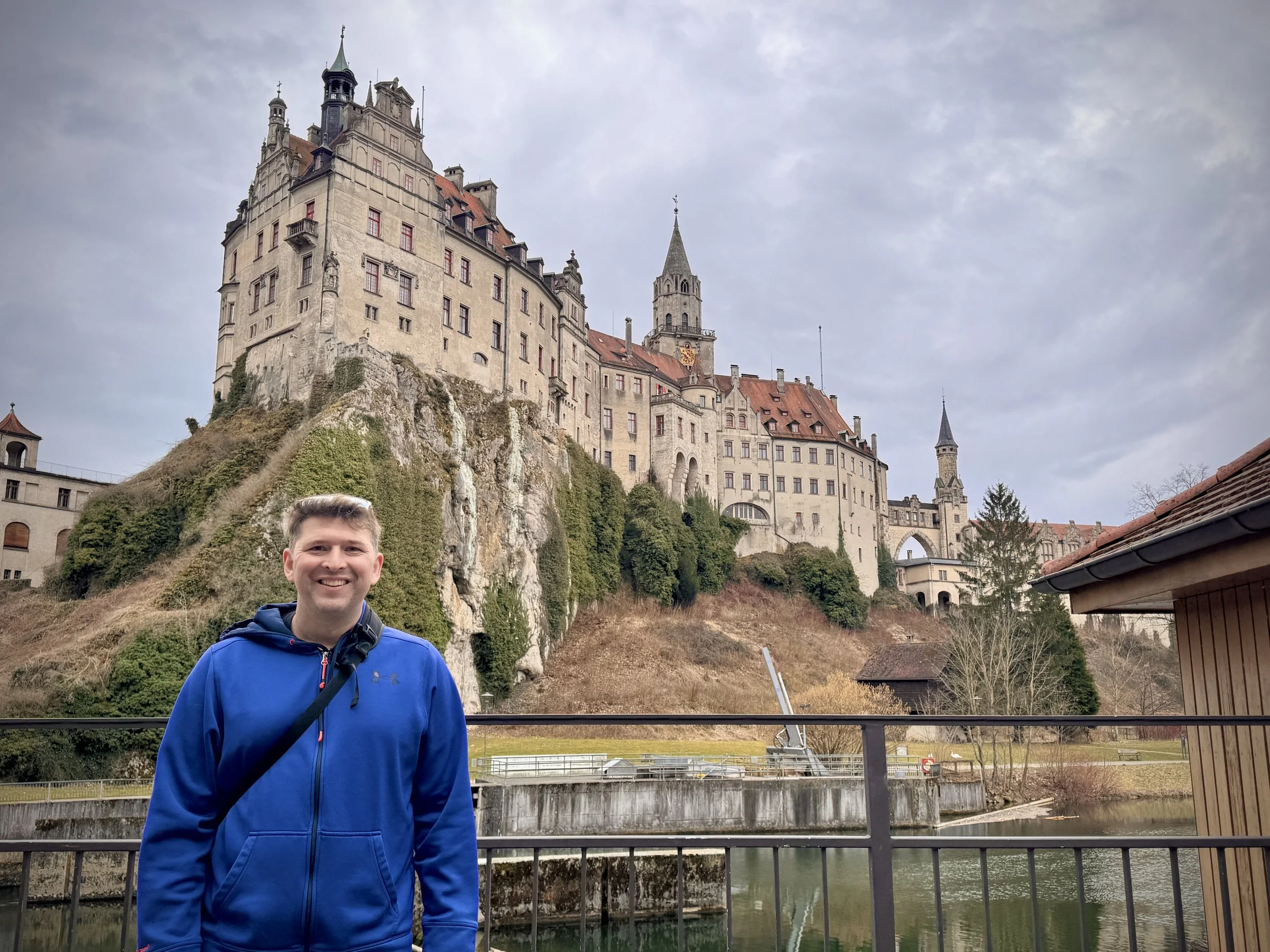 A man standing in front of Hohenzollern Castle in Sigmaringen, Germany, during winter, with the historic hilltop castle rising above the Danube River on an overcast day.