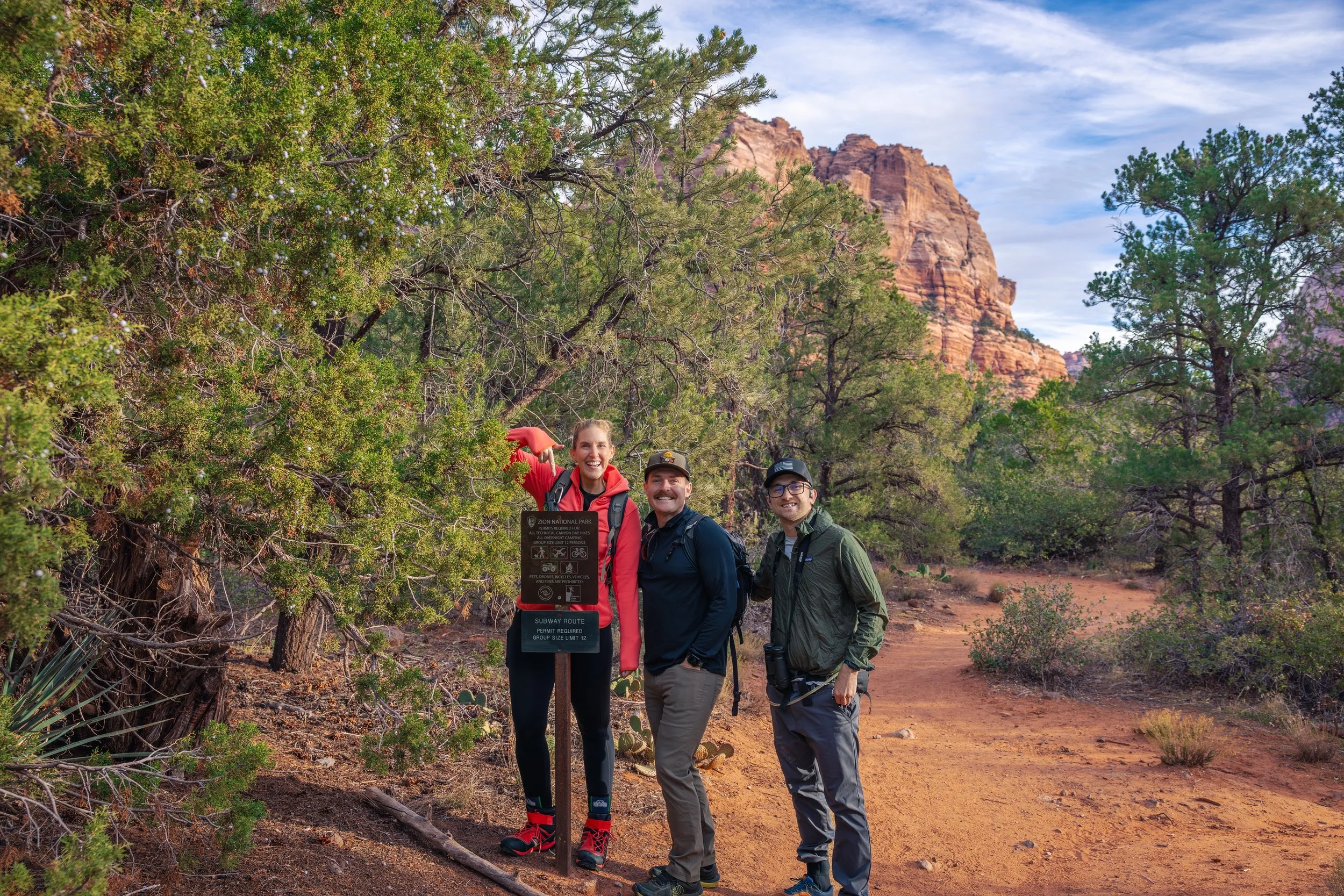 Three hikers standing beside a Subway Route trail sign in Zion National Park, Utah, with red rock cliffs and desert vegetation surrounding the trail.