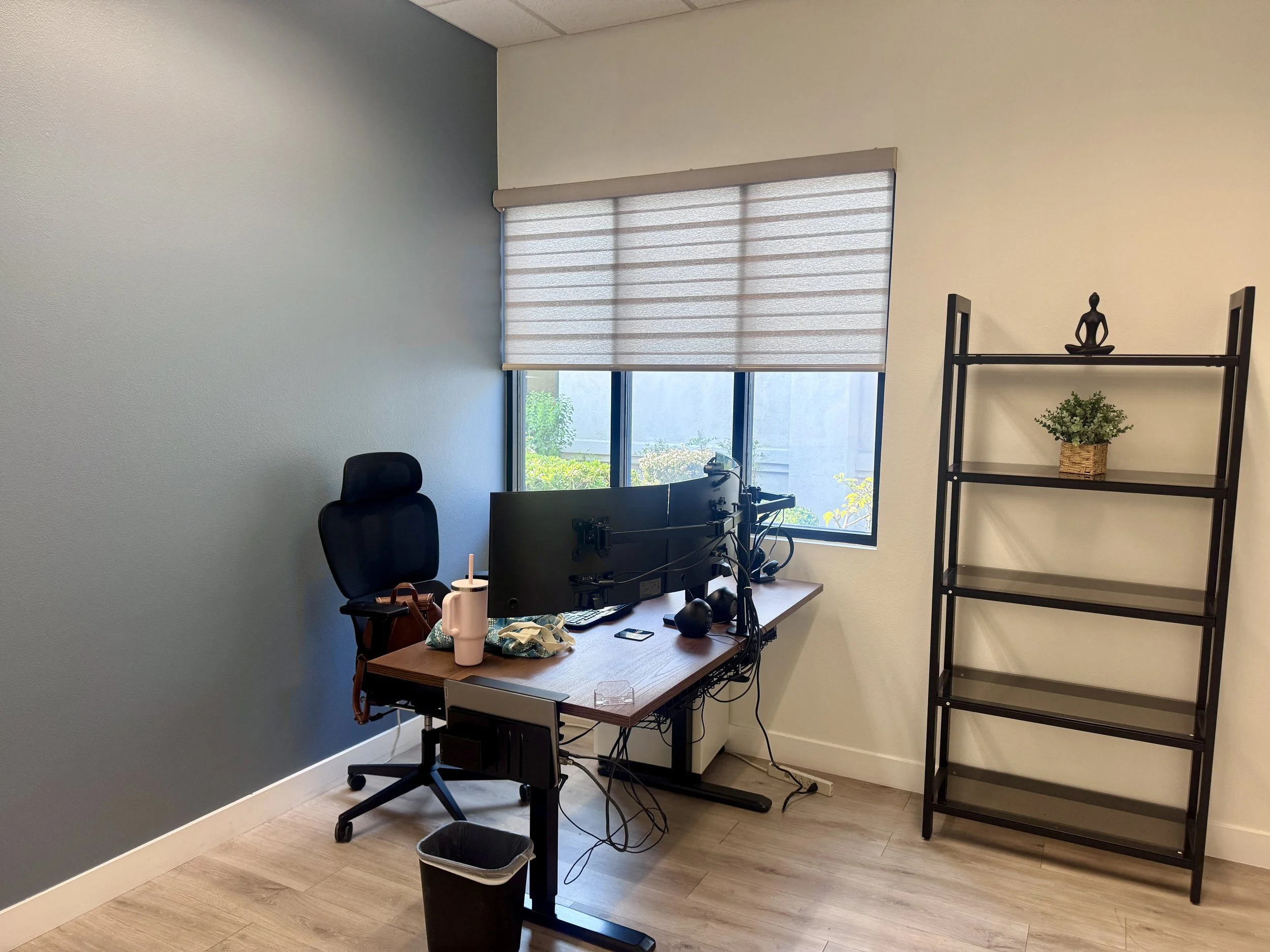 A minimalist office with a standing desk, dual monitors, office chair, and shelving unit, set up in a bright room with a window and neutral decor.