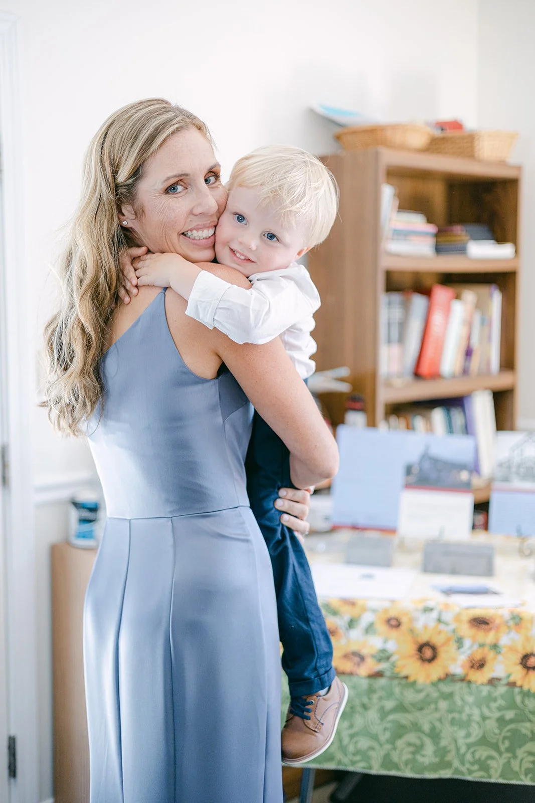 A woman holding a young boy in an indoor setting with bookshelves and a table in the background.