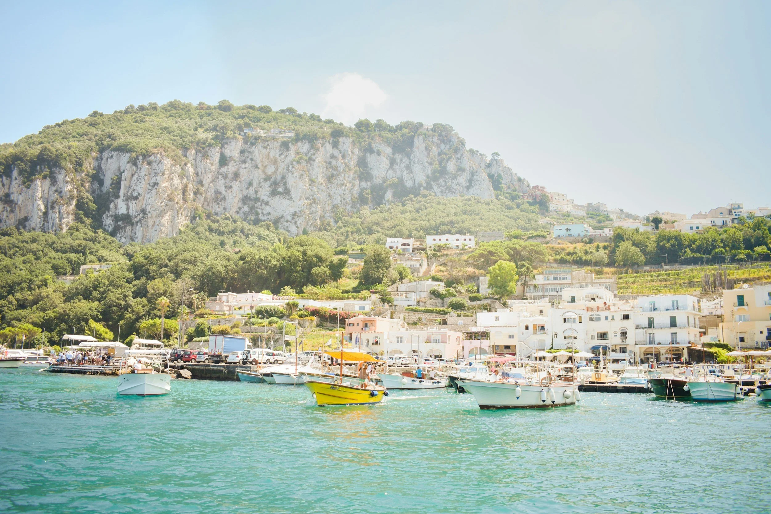 Boats docked at a harbor with a hillside town and rocky cliffs in the background.