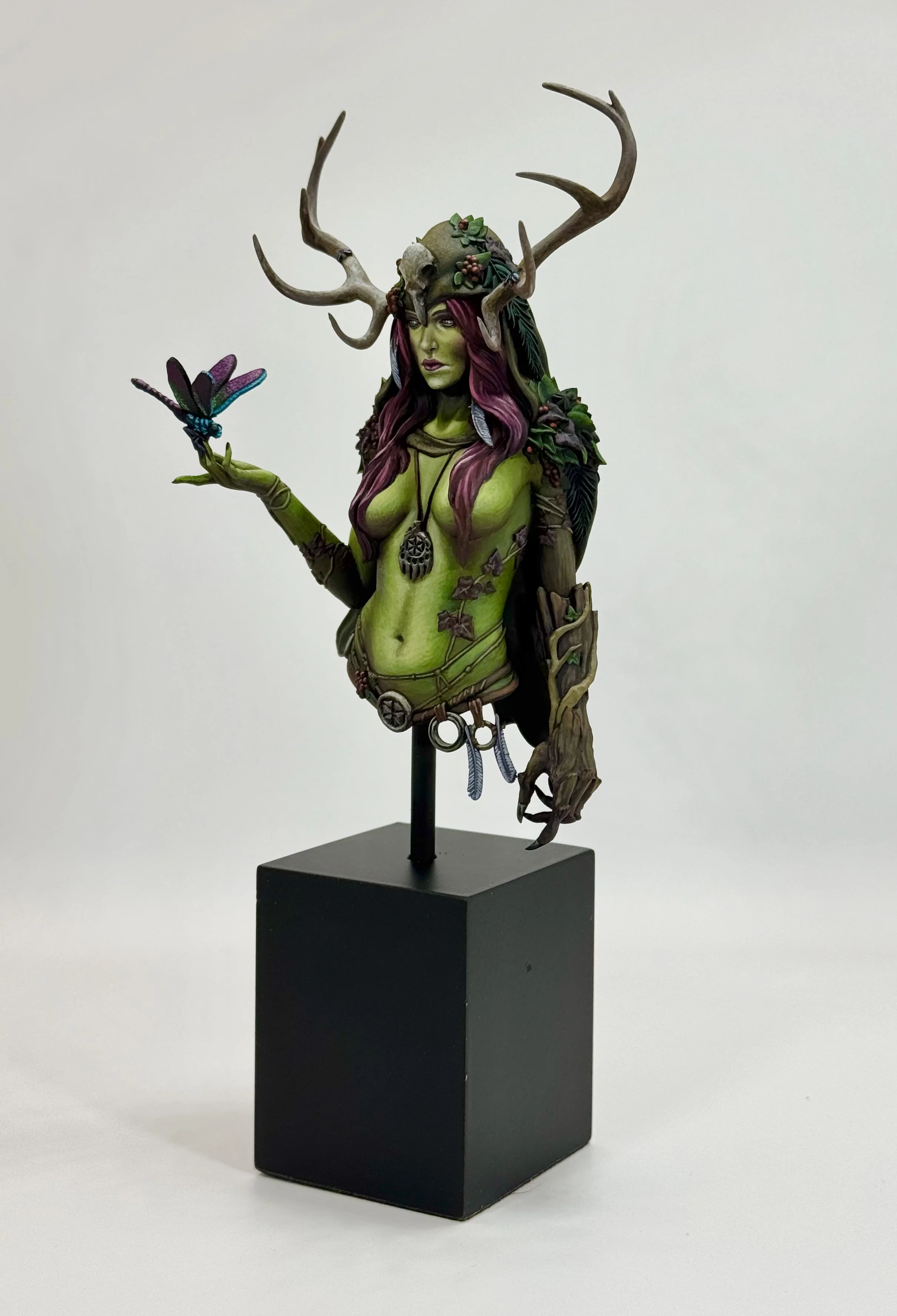 A fantasy sculpture of a female figure with green skin, long purple hair, and deer antlers, holding a colorful butterfly in her hand, adorned with leaves, skulls, and jewelry, standing on a black pedestal.