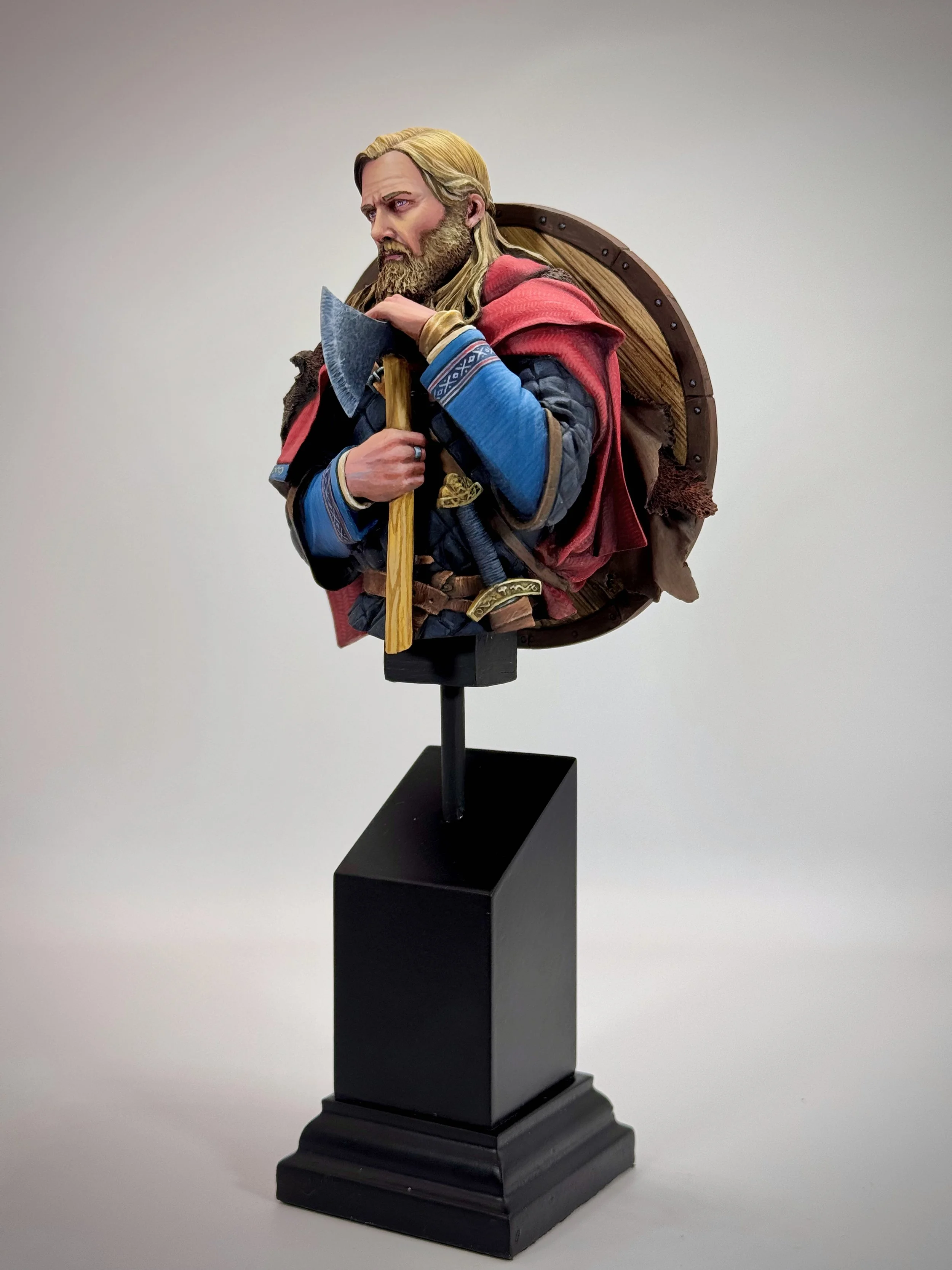 Wax figure of a medieval warrior with long blonde hair, beard, wearing a blue and red outfit, holding an axe, with a shield strapped to his back, standing on a black pedestal.