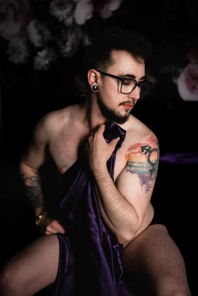 Man with a rainbow tattoo on his arm is sitting down and holding a sheet over him while posing for a boudoir photo in Vancouver, Washington.