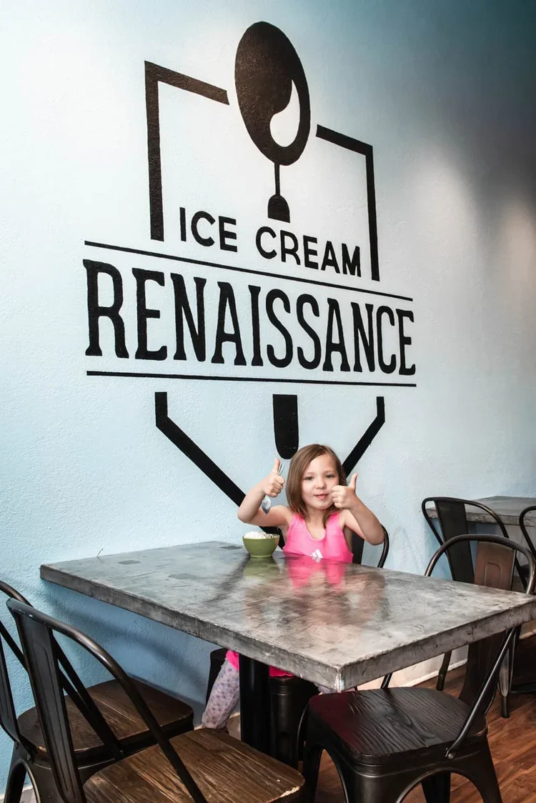 A little girl is sitting down at a table enjoying ice cream and holding two thumbs up. She is sitting in front of a logo that reads "Ice Cream Renaissance."