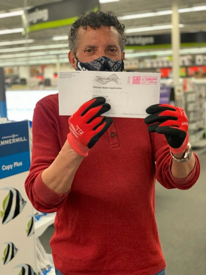 Tim Stevens holding up a mail-in ballot application while wearing gloves and a mask.