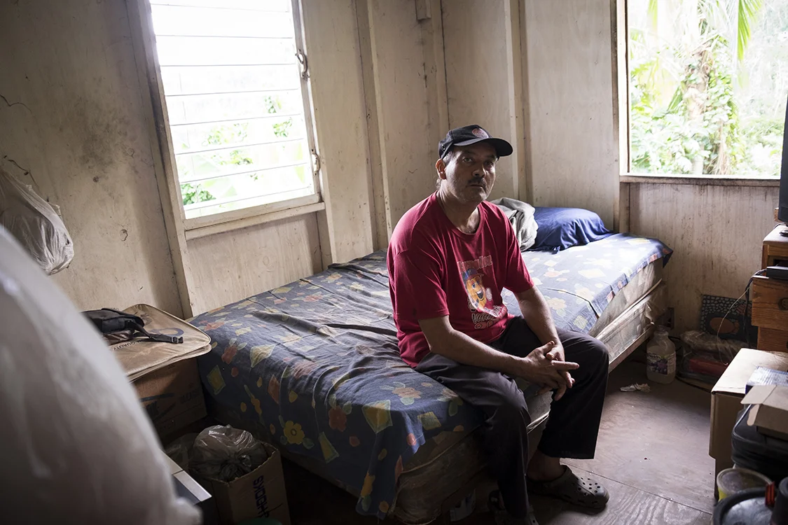A man sits on his mold infested bed. He is a poor man, and knows no one is going to come to help him fix his home; so, he plans to do all the work himself.