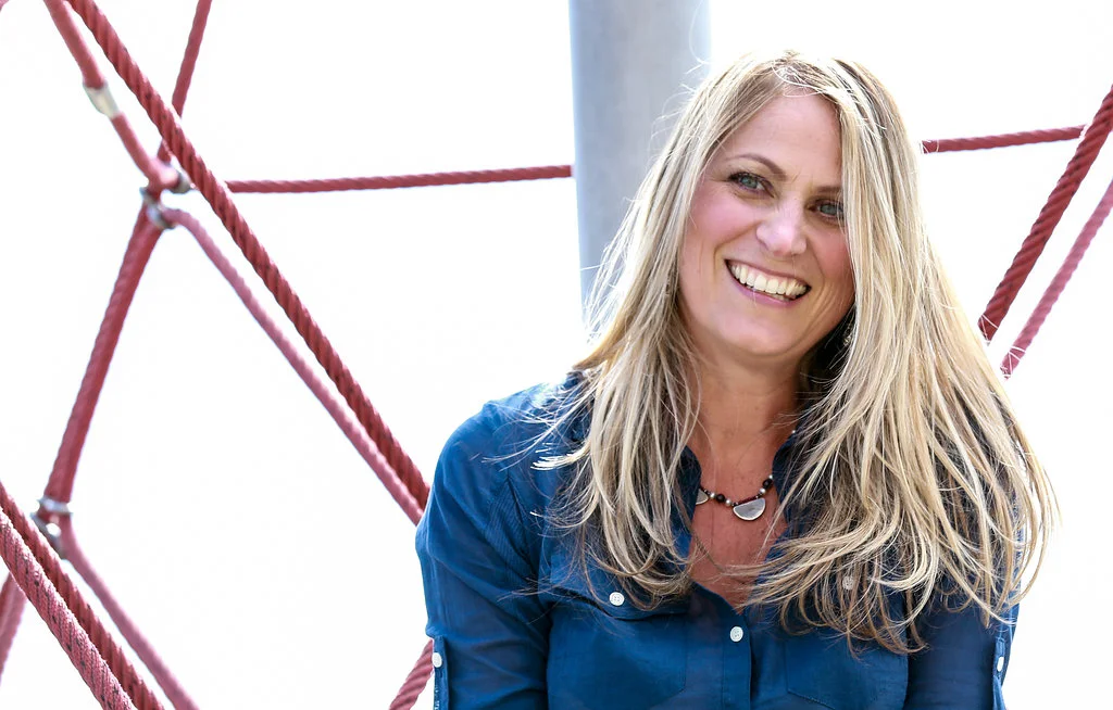 A woman with long blond hair smiling outdoors, sitting near red ropes on a playground or climbing structure.