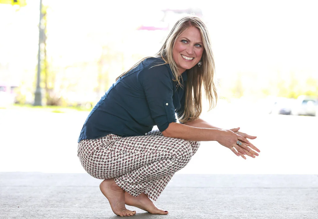 A smiling woman with long blonde hair, wearing a dark blue shirt and patterned pants, squatting barefoot outdoors on a concrete surface.