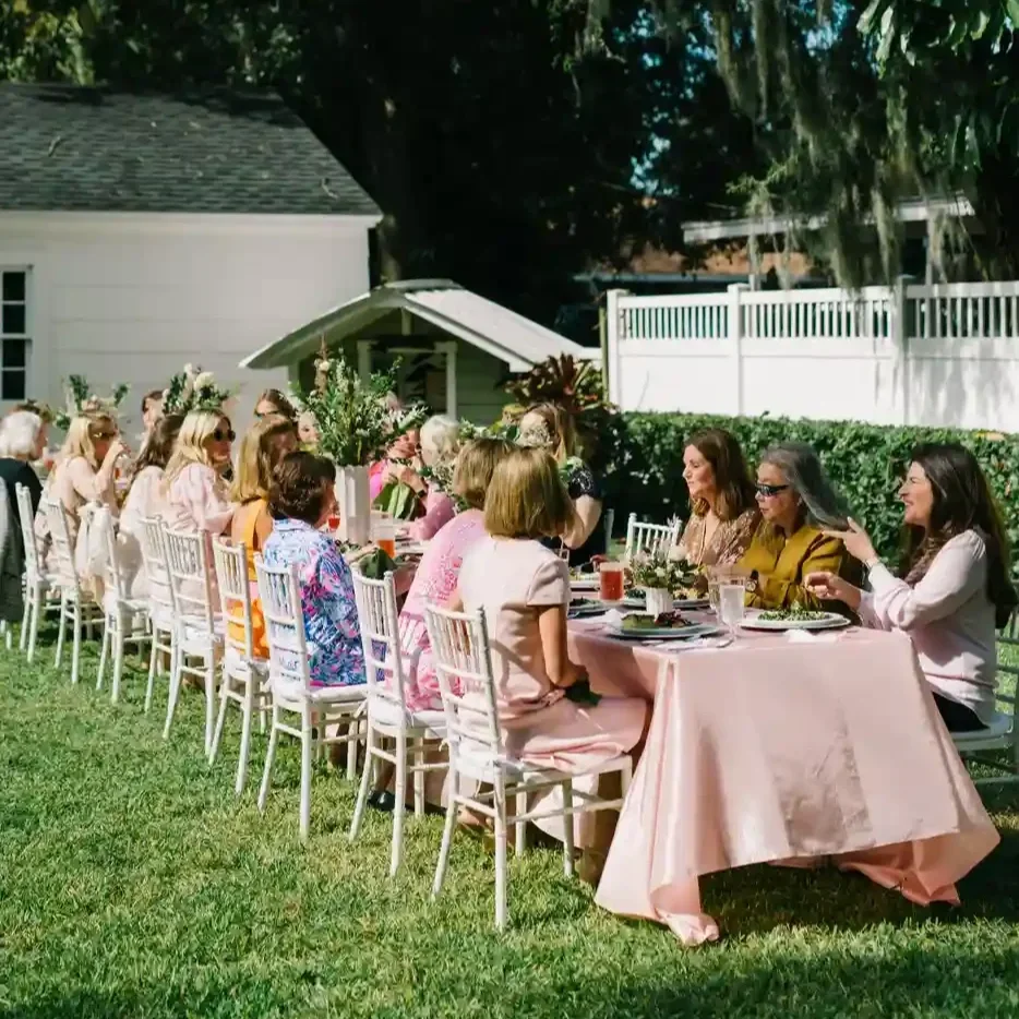 long table covered with a pink tablecloth outside surrounded by women sitting and eating at a baby shower