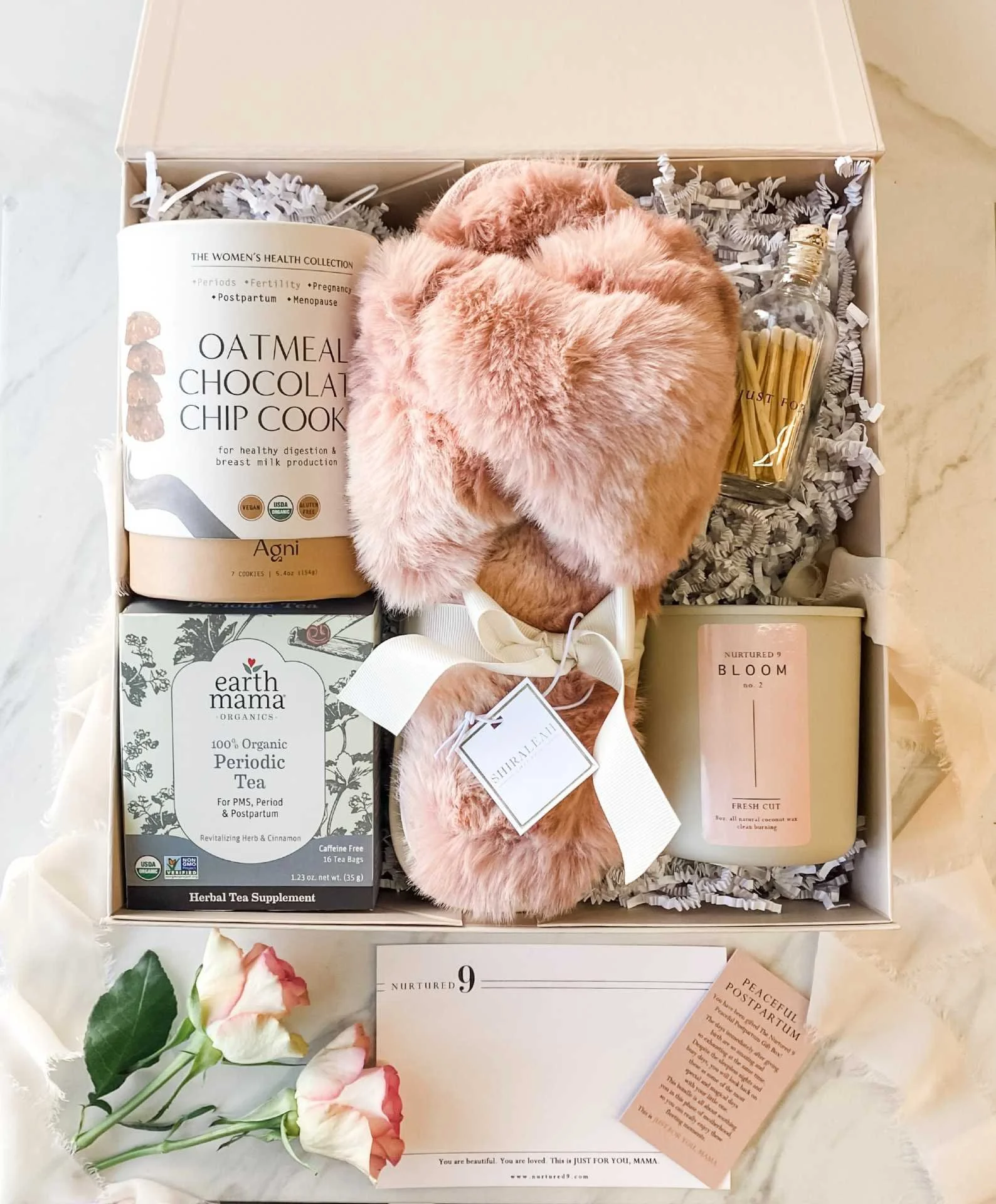 A postpartum gift box with pink fluffy slippers, a candle, herbal tea, a jar of apothecary matches, a pink card, and pink and white flowers on a marble surface.