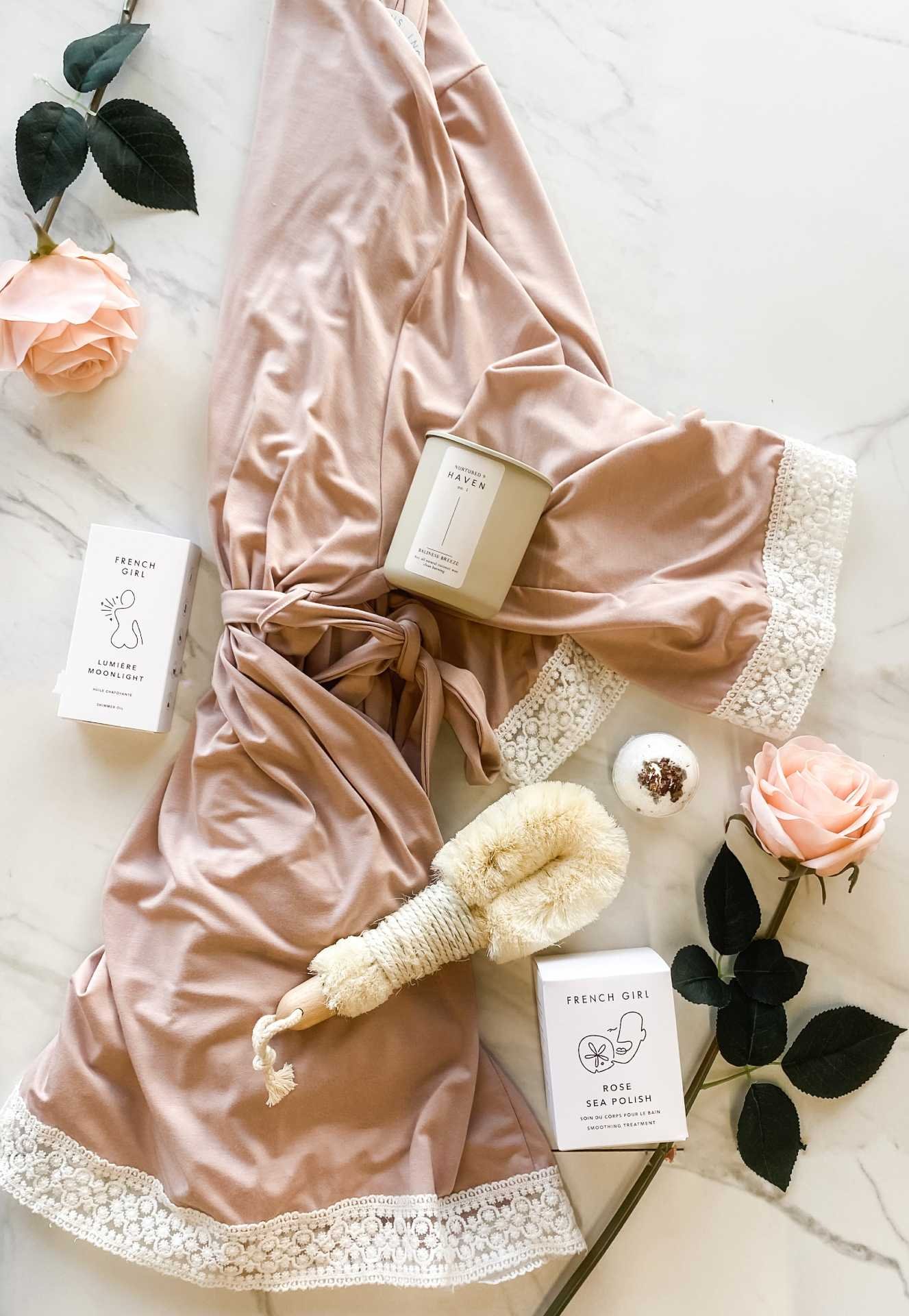 A flat lay of skincare and self-care items on a marble surface, including a pink maternity robe with lace trim, pink roses, a dry brush, a candle, a bath bomb, and skincare products in white boxes.
