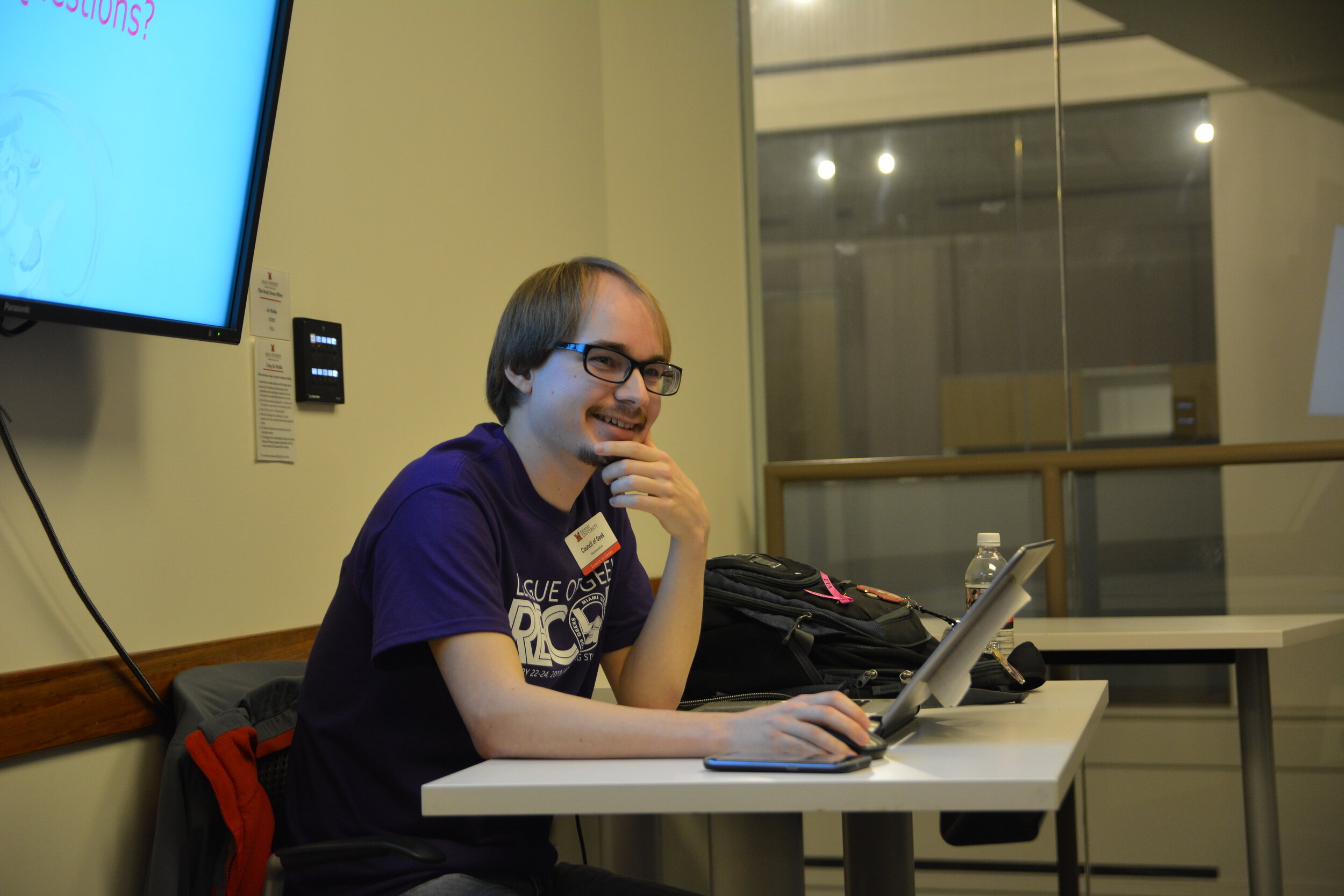 A person in a RECON team shirt sitting in front of a computer on a desk facing the right smiling at something off camera.