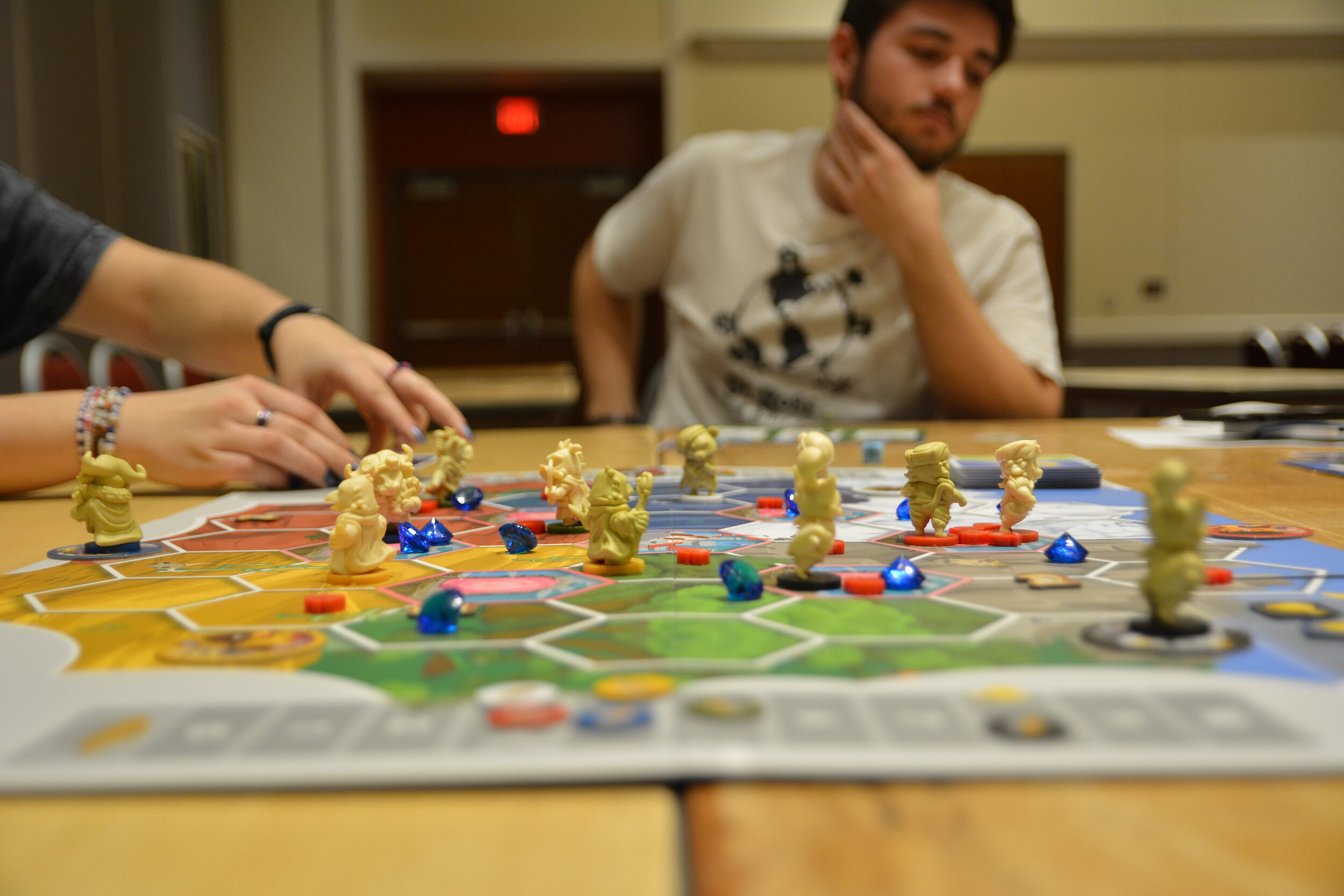 A close up of a board game with somehands on the left and behind the board game facing the camera is a blurry person.