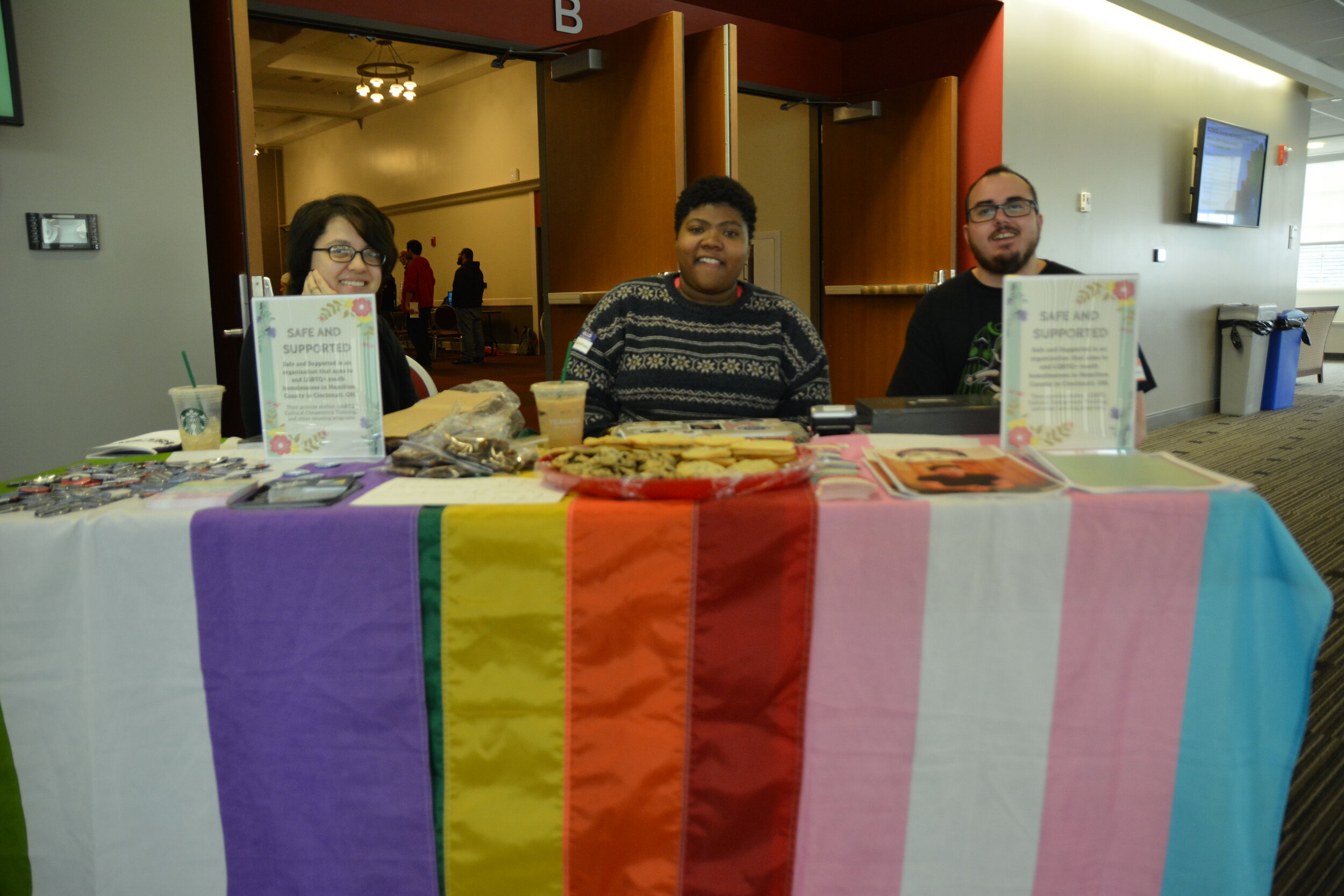 3 people sitting in front of the Pavilion entrance at a table with pride flags as tableclothes facing and smiling at the camera 