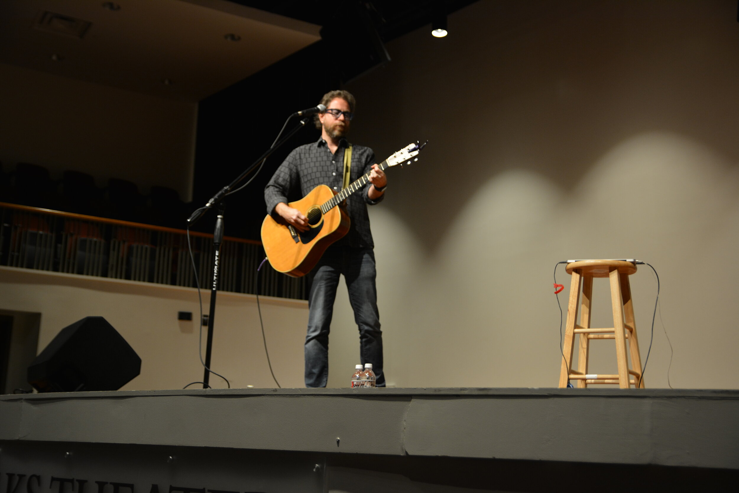 Jonathon Coutlon on stage with his guitar, a mike, and a stool
