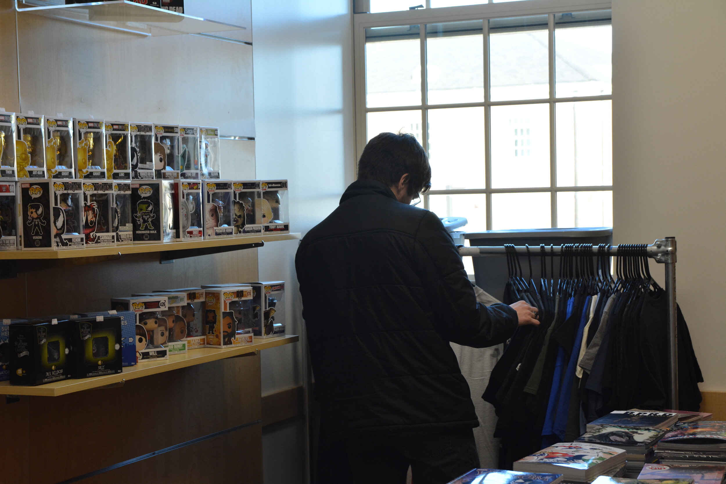 Someone in a dark coat's back looking at some shirts in front of a window next to comics and funk pops in the room in Armstrong that Future Great Comics rented out to sell things.