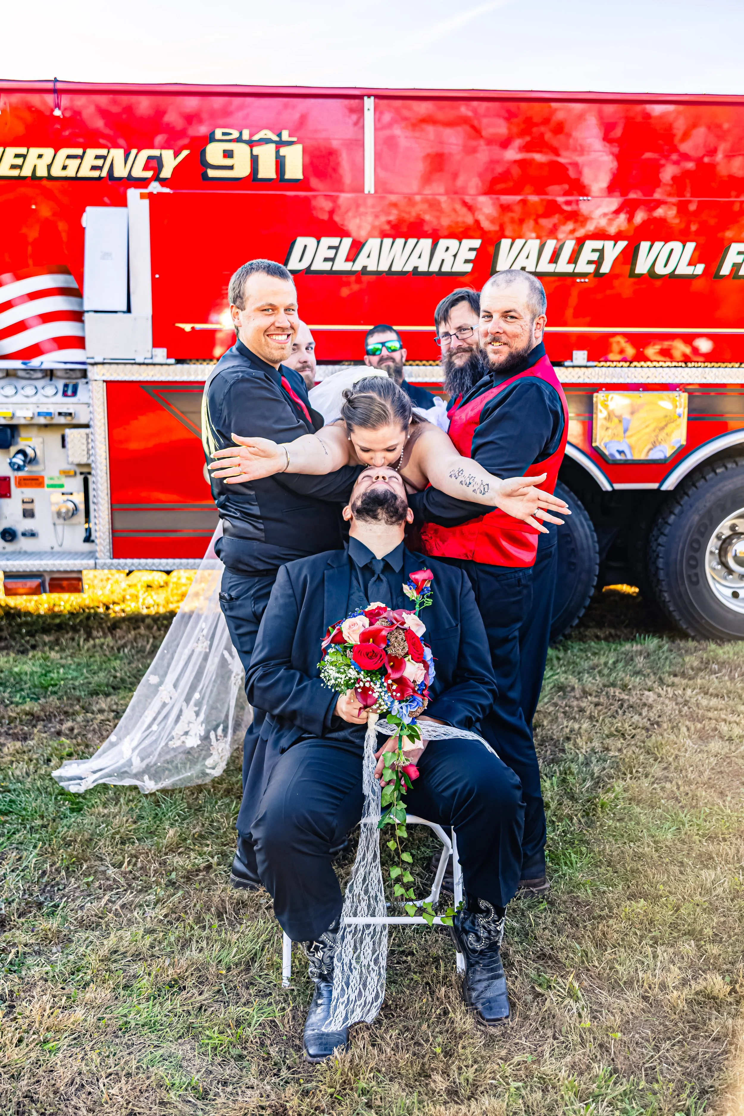 Group of people with tuxedos and wedding dress, one person sitting with a flower bouquet, others hugging, in front of a fire truck labeled Delaware Valley Vol. Rescue.