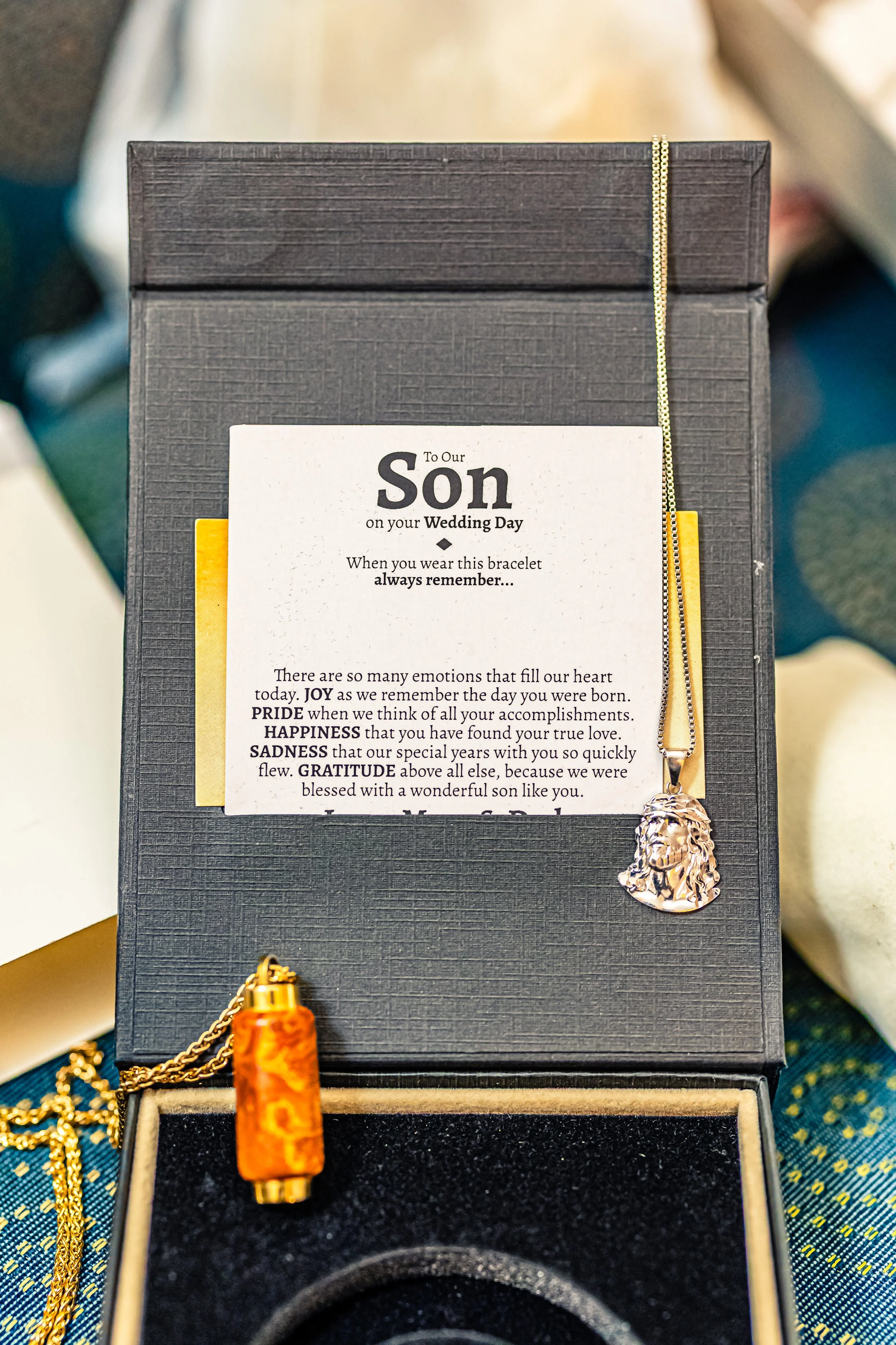 A jewelry gift box containing a lion head pendant necklace, a small orange and gold vial-shaped pendant, and a printed note for a son to remember on his wedding day, inside a gift box.