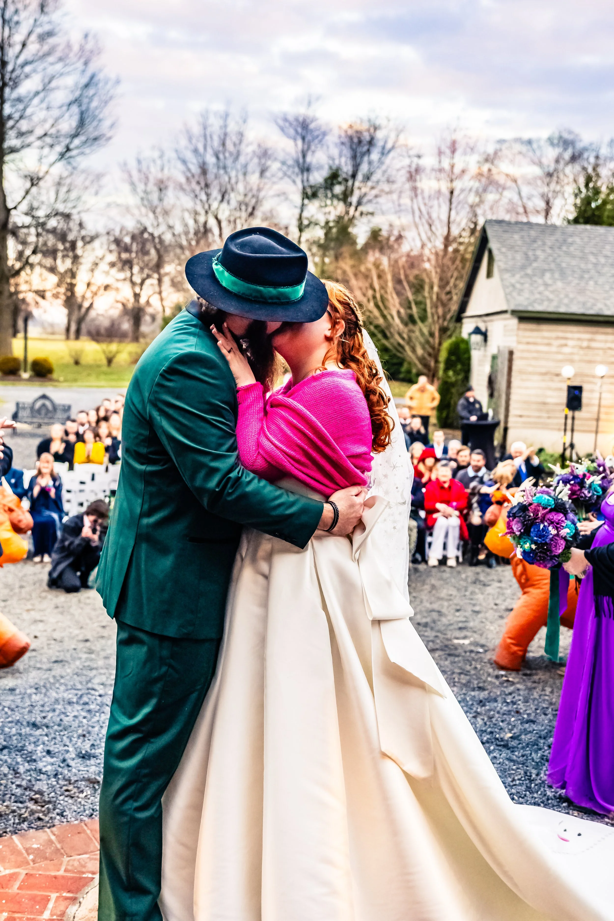 A wedding ceremony outside, showing a couple kissing with the bride in a white gown and the groom in a green suit and black hat with a green band. Guests are seated in the background, and the scene takes place during early evening or sunset.