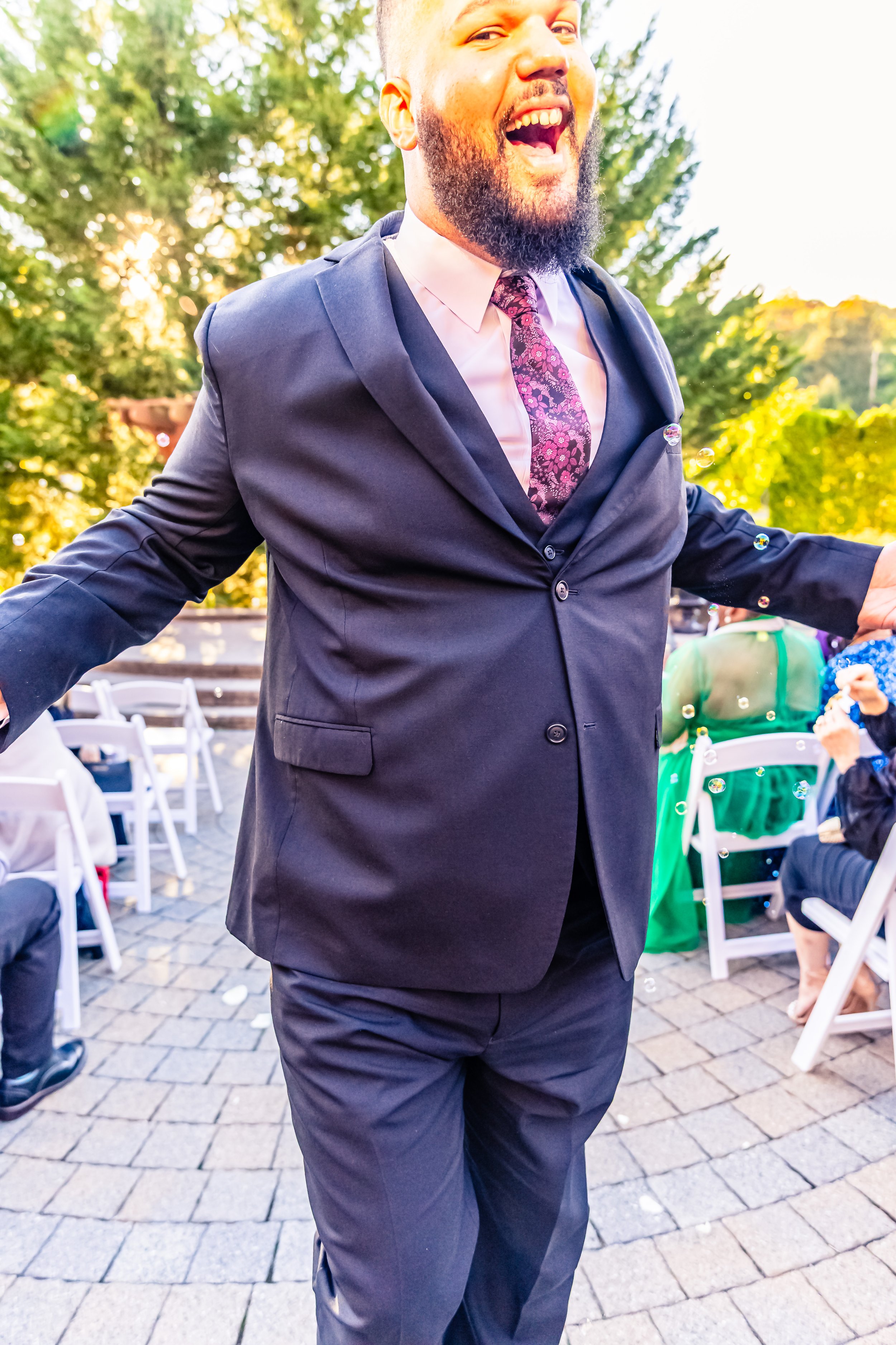 Happy man in a suit celebrating at an outdoor event with trees and decorated tables.