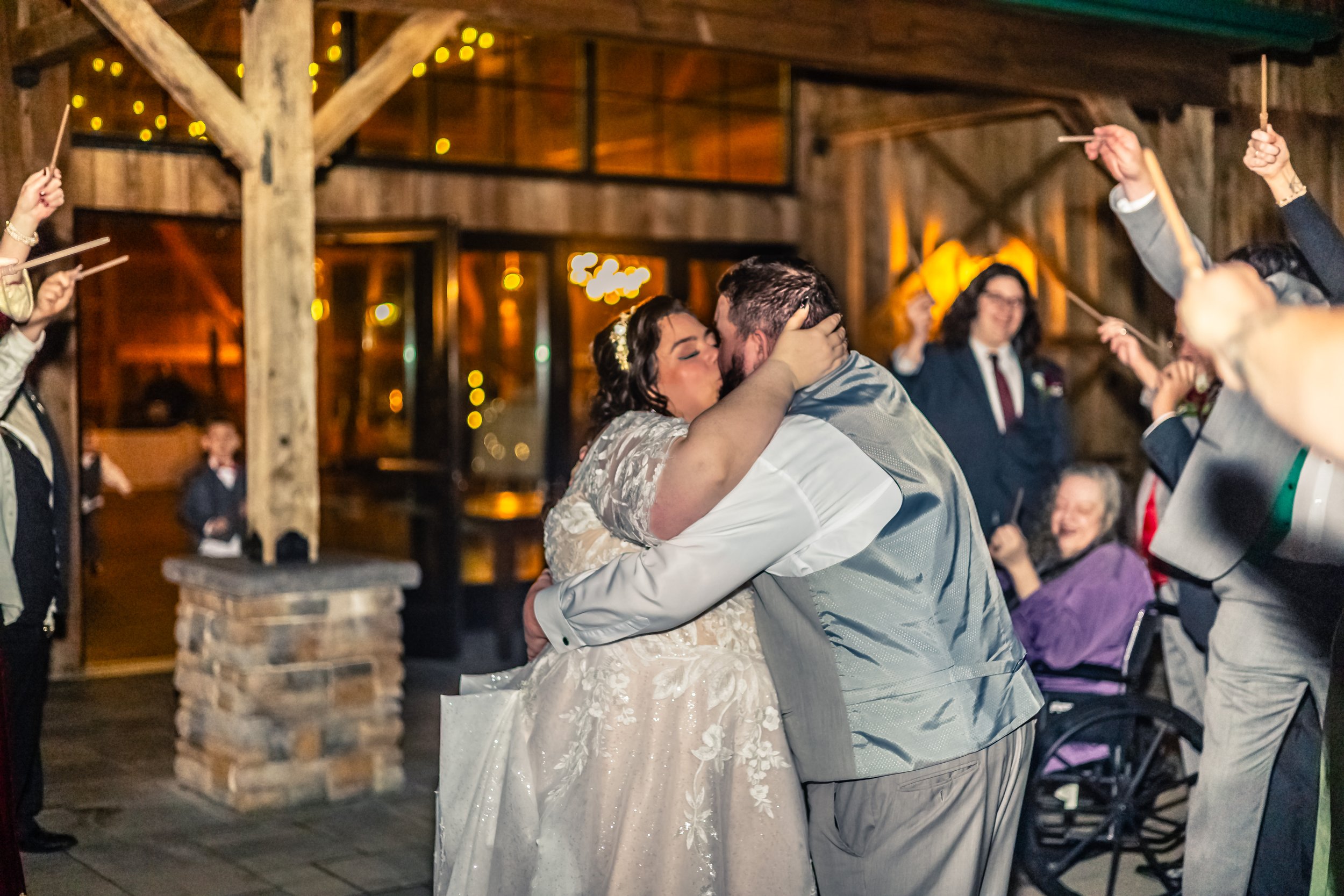 A bride and groom kiss during their wedding reception, surrounded by guests holding sparklers in a rustic wooden venue with warm lighting.