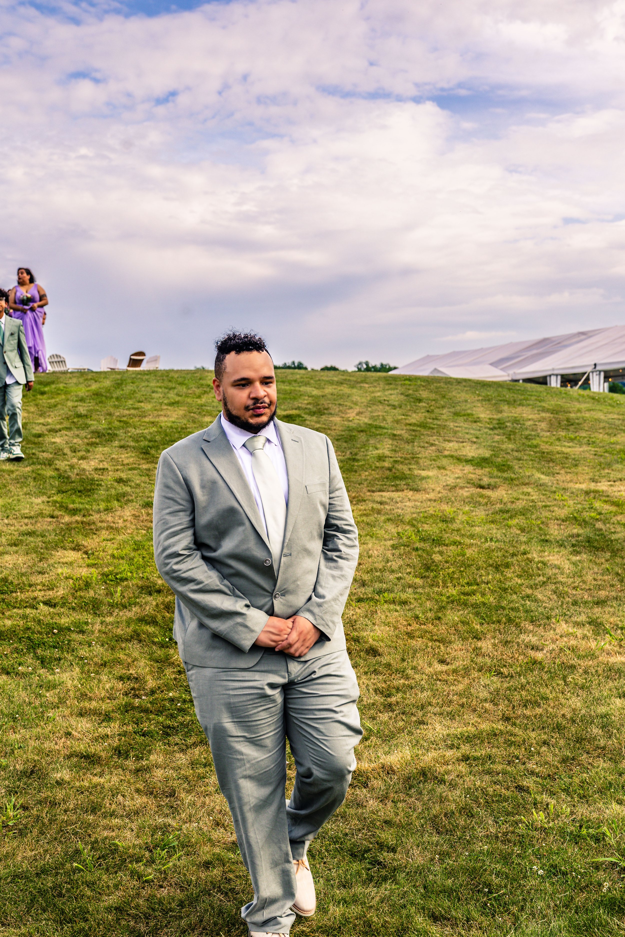 A man in a light gray suit walking on a grassy area outdoors, with a cloudy sky in the background. In the distance, there are two women and another man, some chairs, and a tent.
