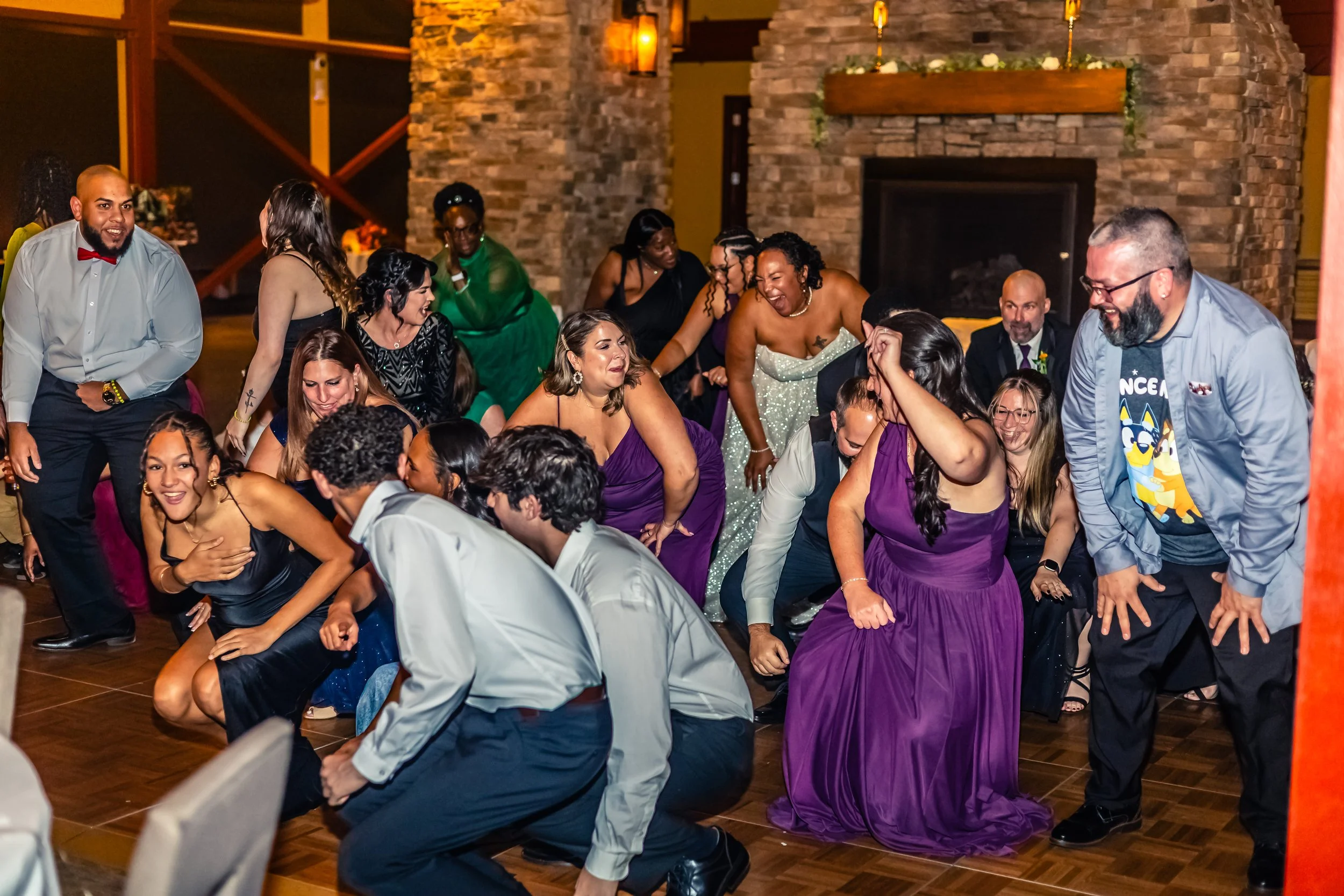 People dancing and having fun at a lively indoor celebration, likely a wedding reception, with a brick fireplace in the background.