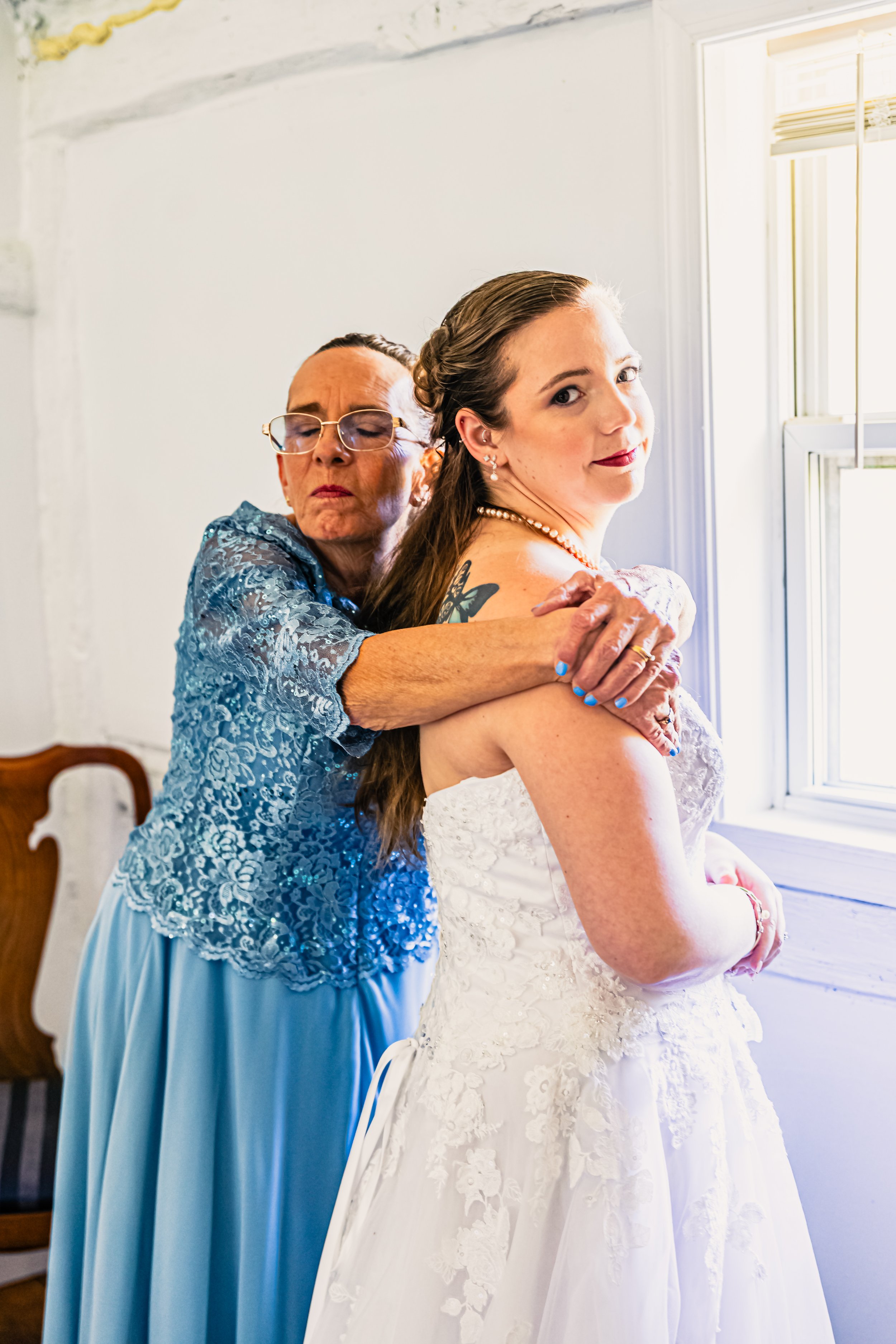 An elderly woman in a blue lace dress embracing a young woman in a white wedding gown near a window, both with a calm and affectionate expression.