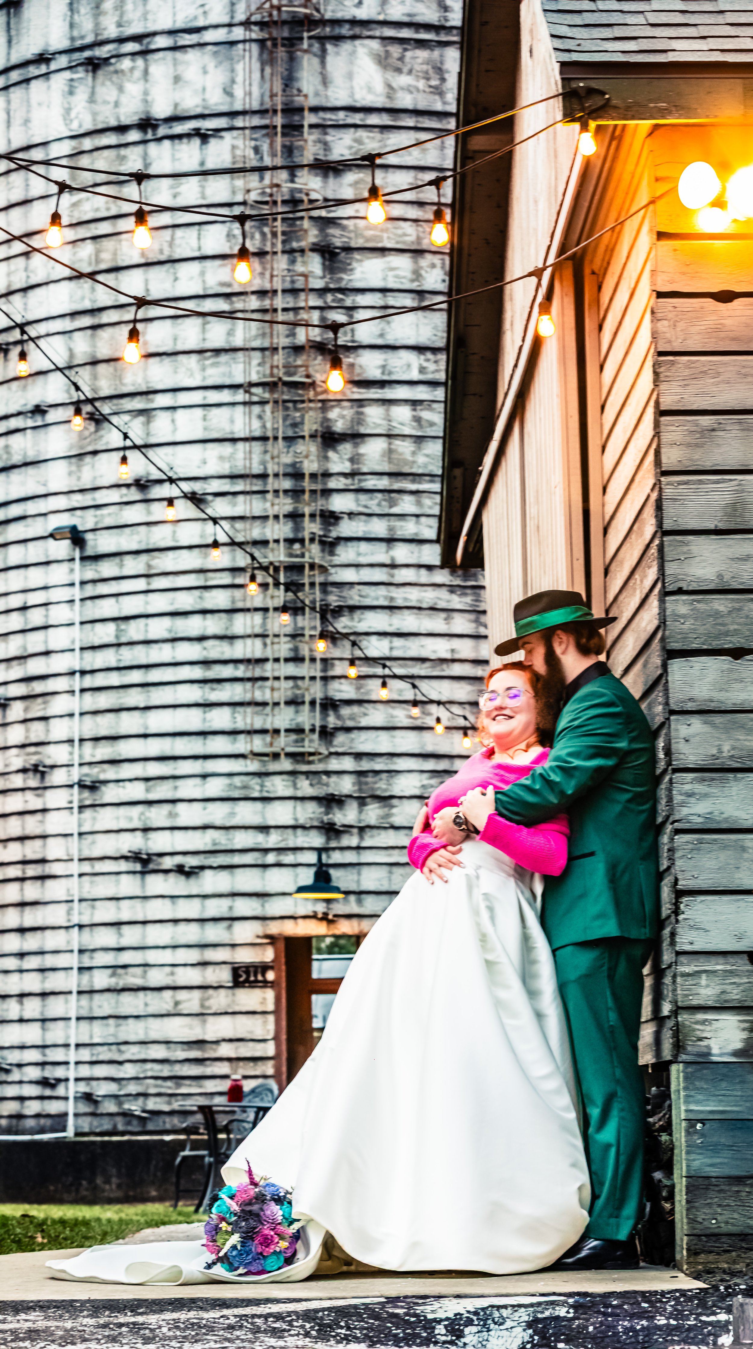 A couple in wedding attire sharing a romantic moment outside a rustic barn with string lights overhead, and a colorful bouquet on the ground.