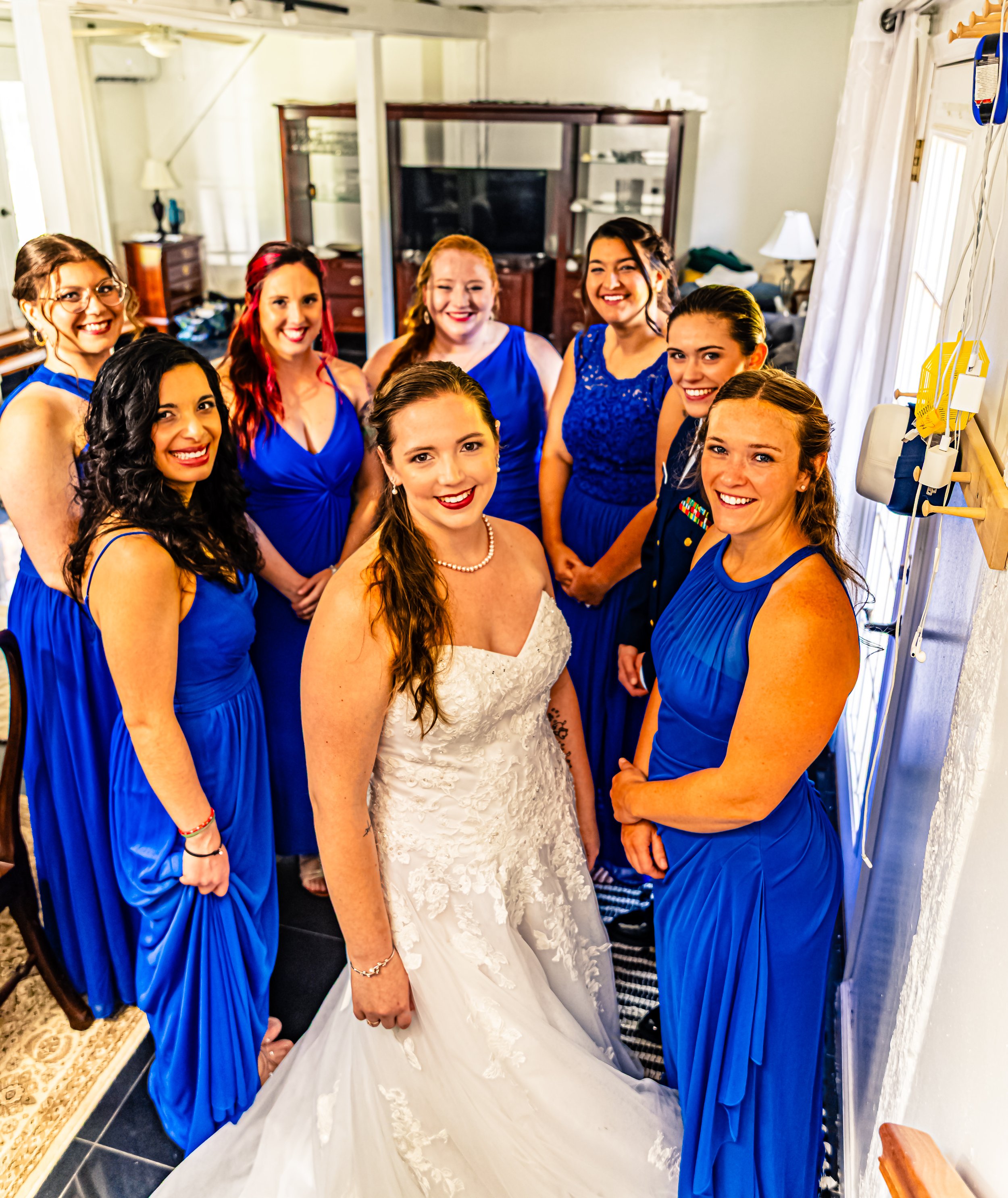 A bride in a white wedding dress surrounded by nine women wearing matching royal blue dresses, all smiling and posing for a photo inside a room.