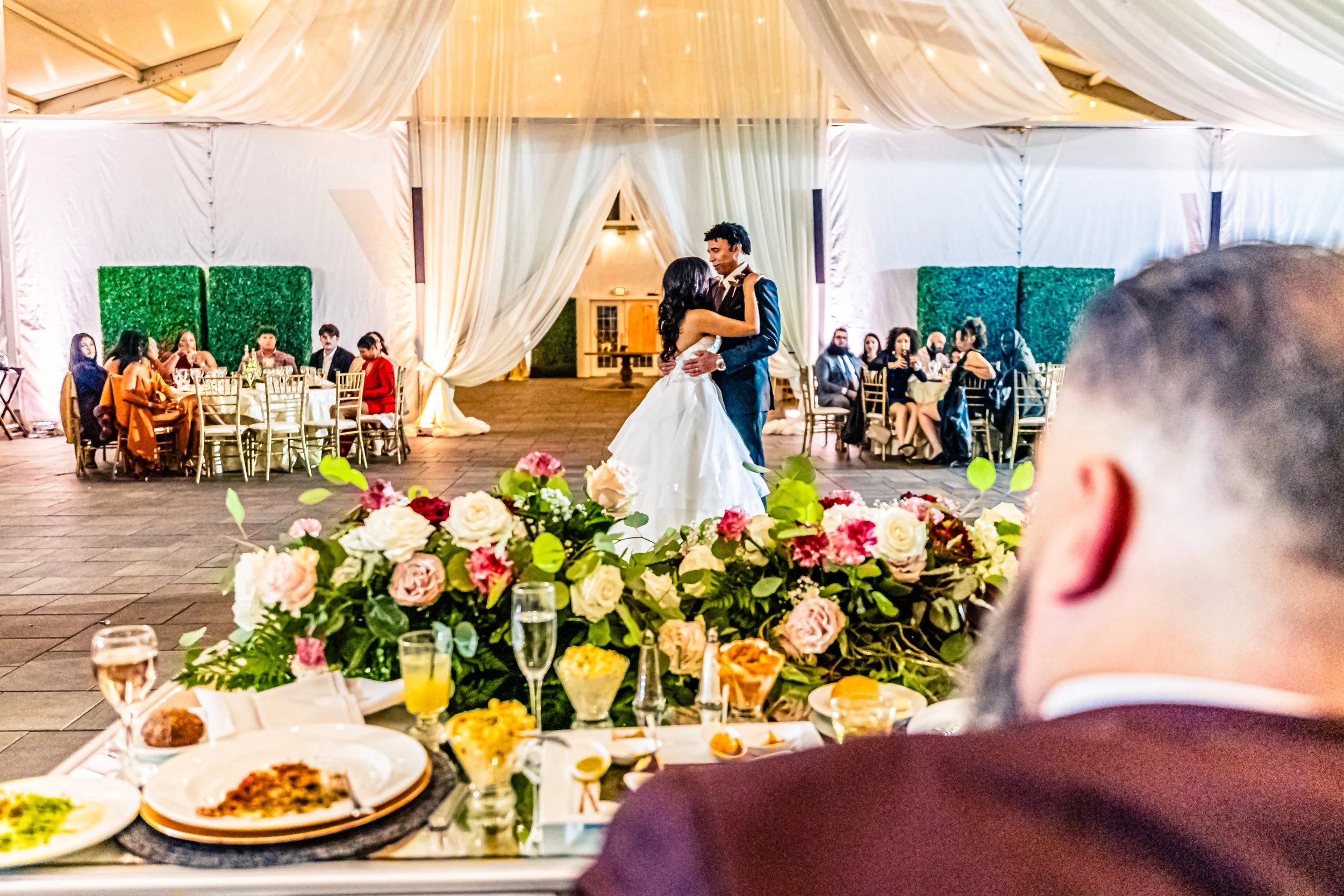 A bride and groom dancing at their wedding reception in a decorated tent, with guests sitting at tables in the background and a floral centerpiece in the foreground.
