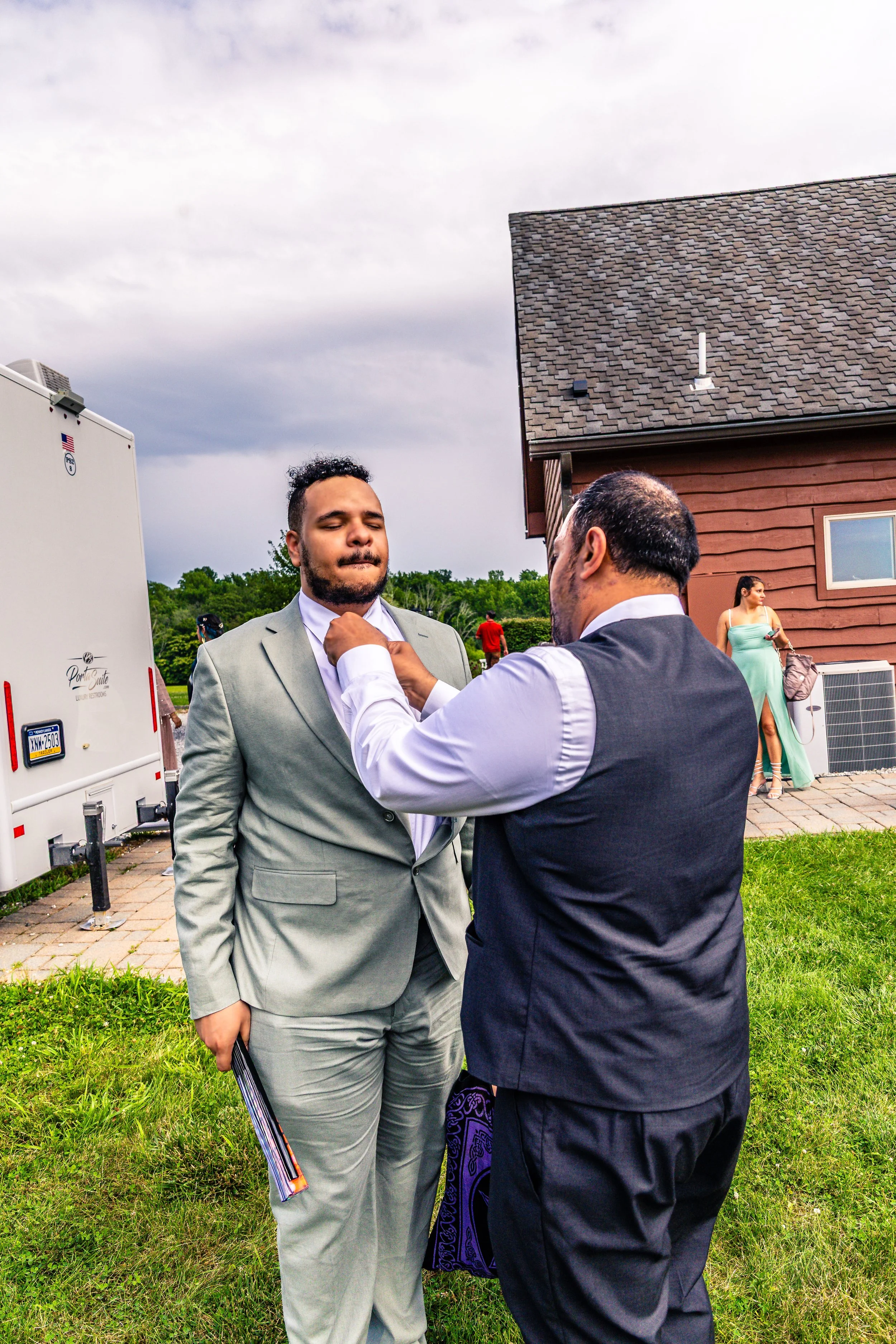 A man in a light gray suit has a boutonniere put on his lapel by another man in a dark vest at an outdoor event, with a woman in a green dress standing nearby.