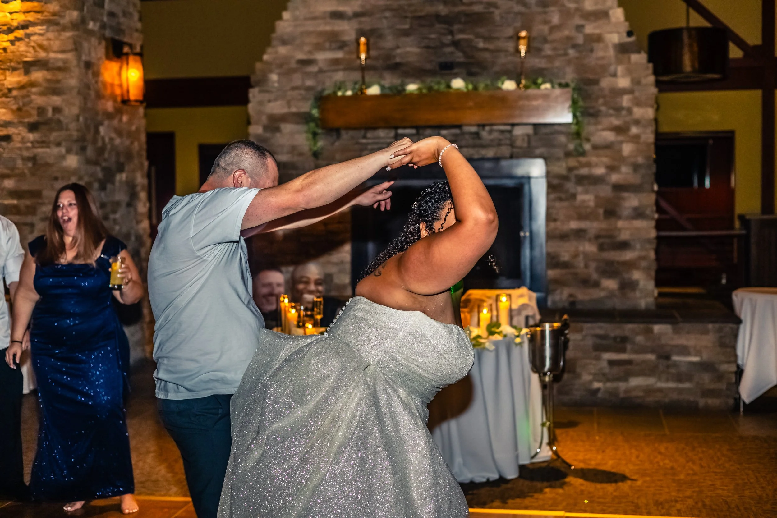 A couple dancing at a wedding reception, with a man twirling a woman in a sparkly dress, surrounded by other guests, a stone fireplace, and candlelit tables.