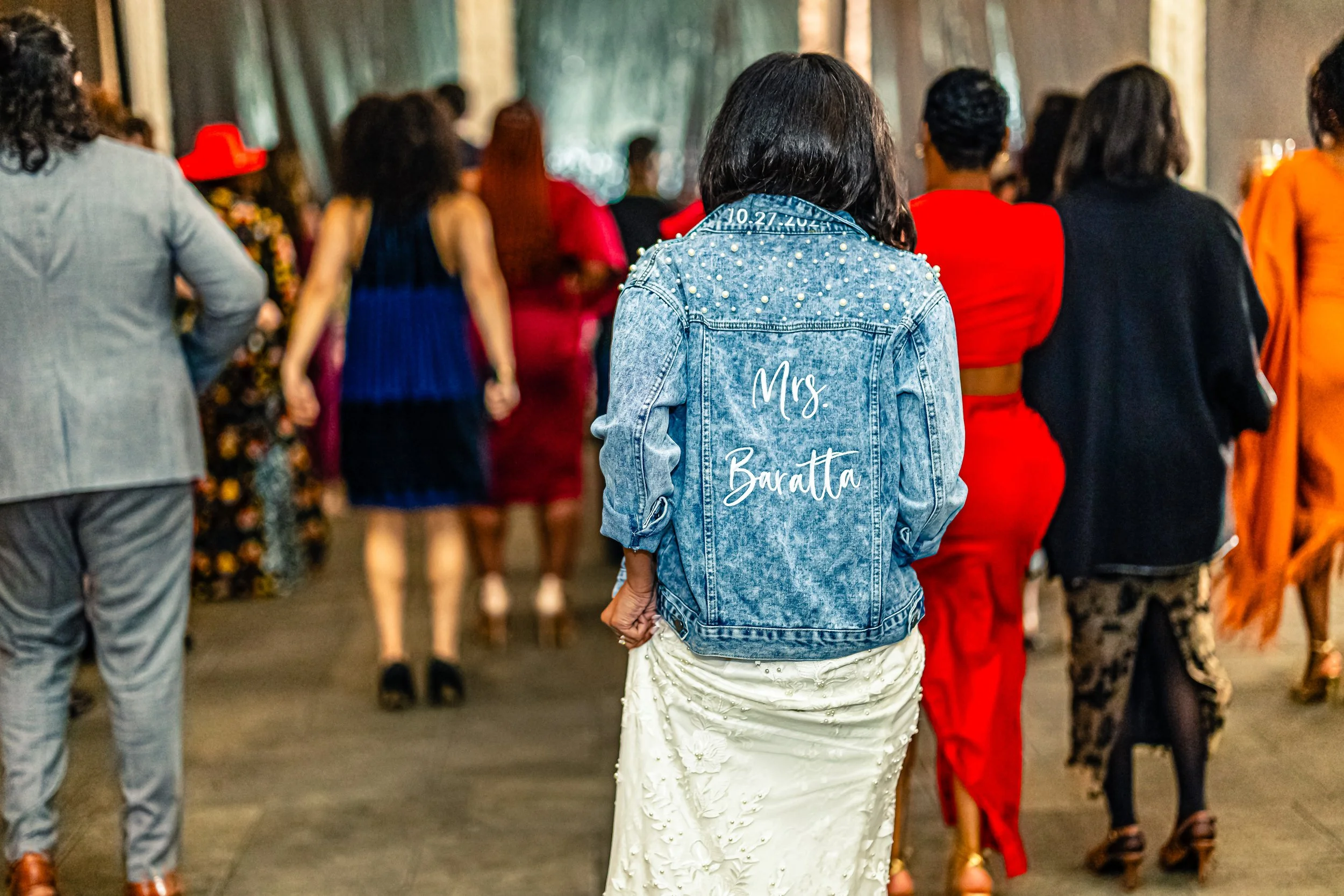 Woman wearing a denim jacket with pearl embellishments and 'Mrs. Baratta' written on the back, at a formal event or celebration with other women in elegant dresses.