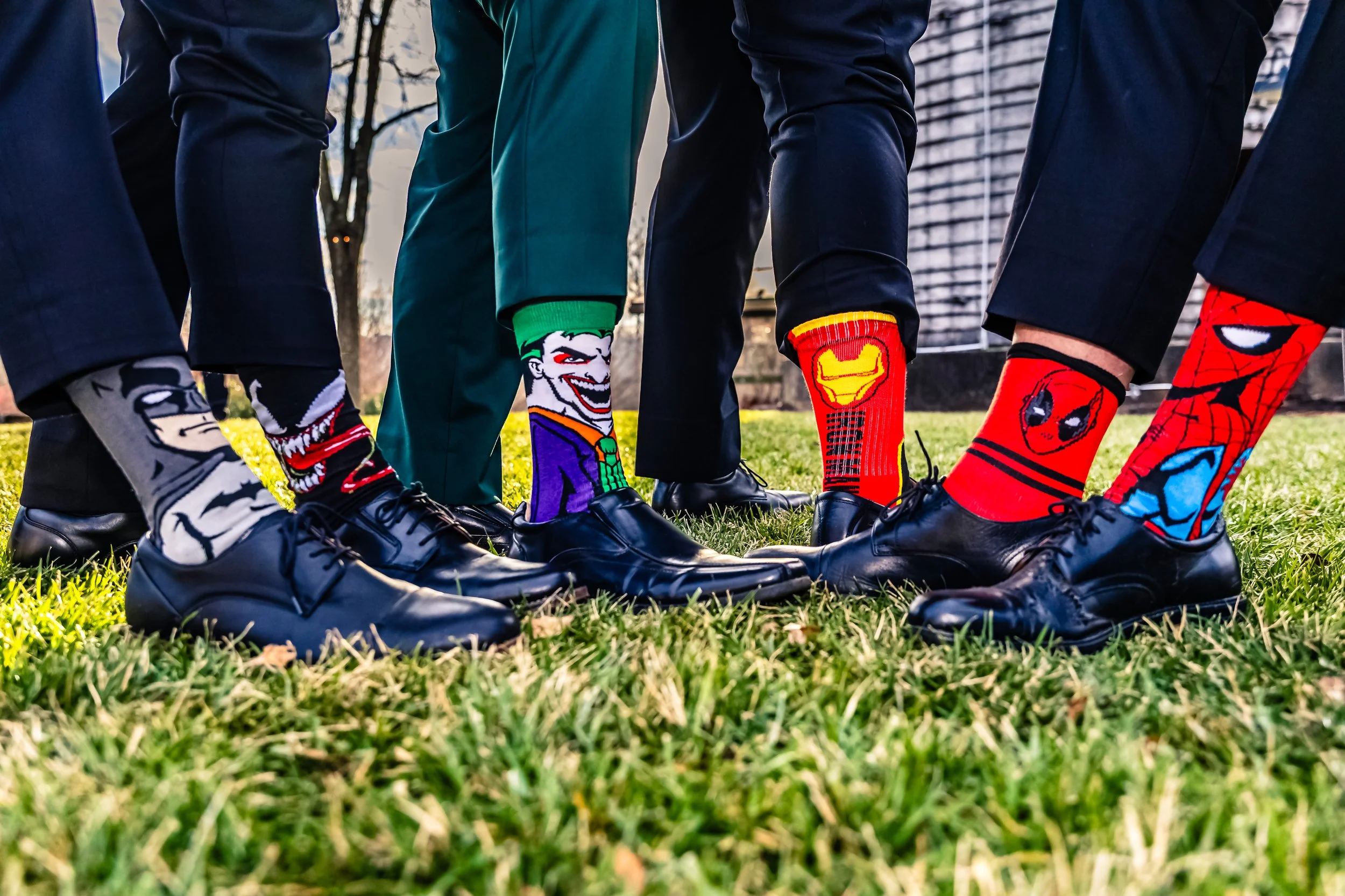 A group of people dressed in business suits standing on grass, showing off colorful comic book character socks and black dress shoes.