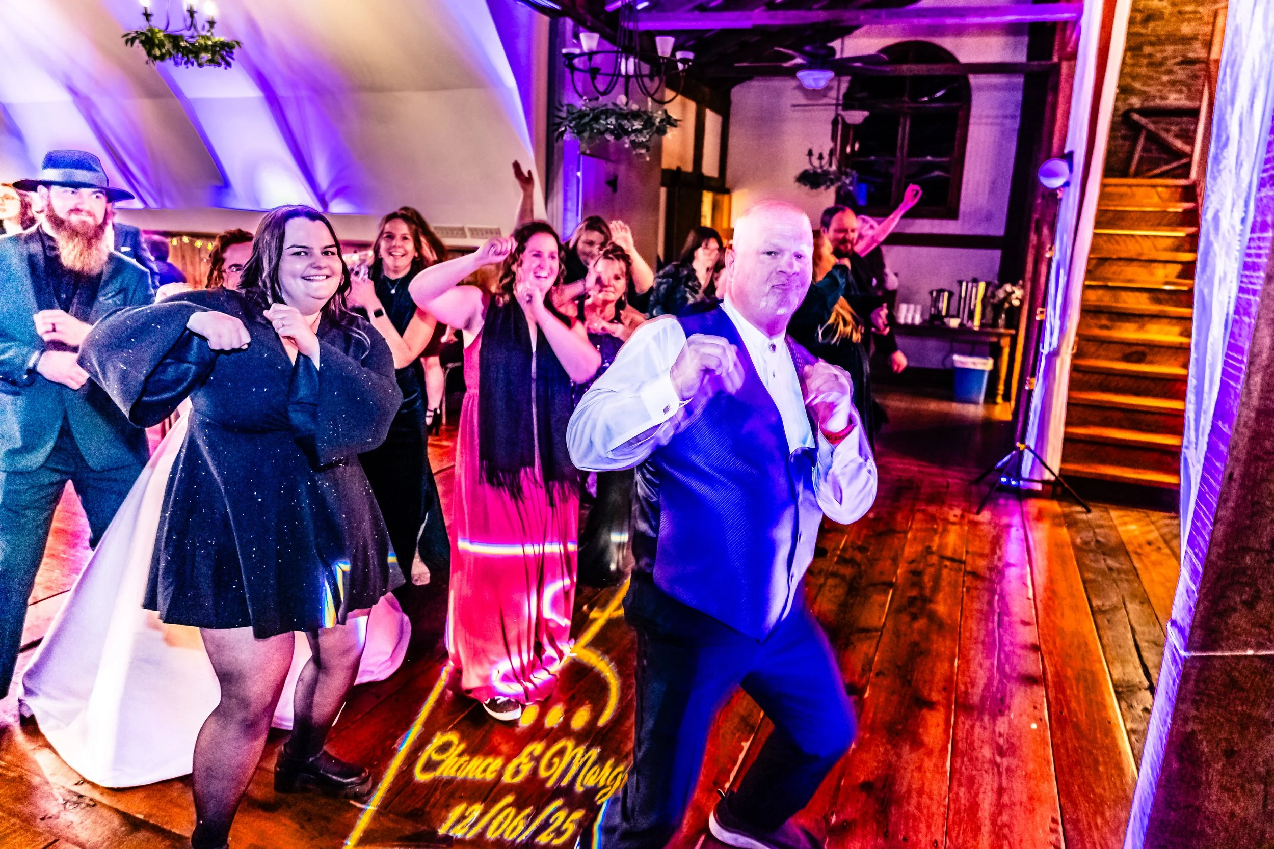 People dancing and celebrating at an indoor party or wedding reception, with a man in front striking a playful pose, amidst colorful lighting and wooden flooring.