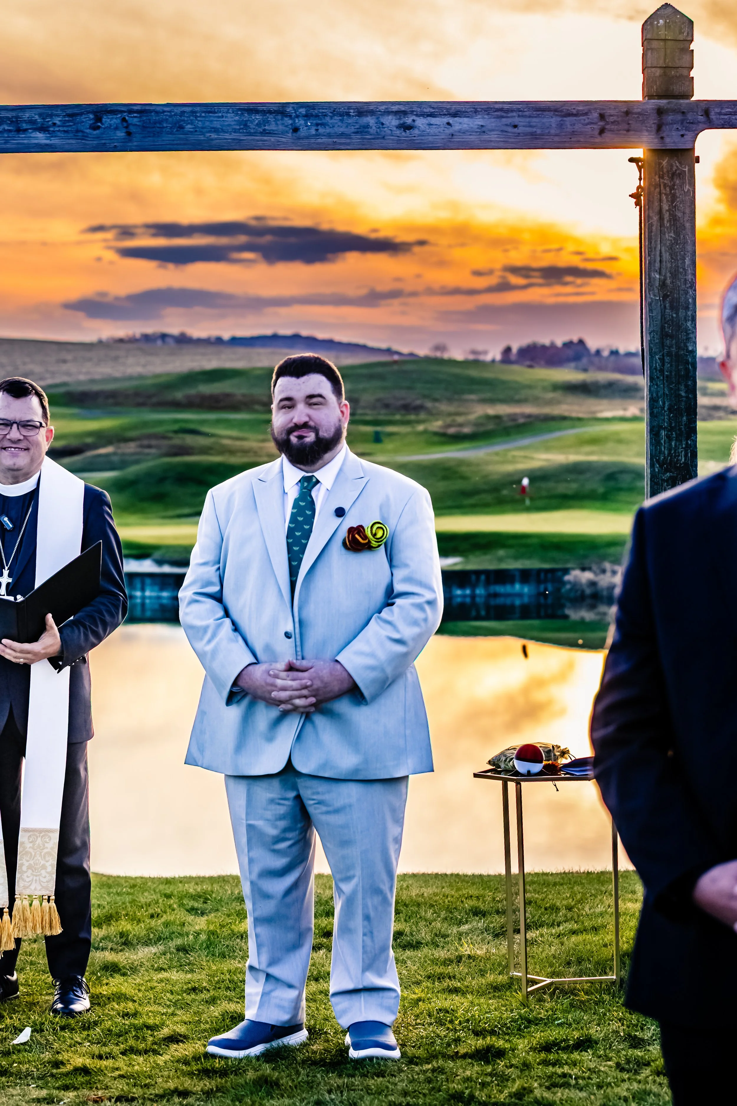 A man dressed in a white suit standing outdoors during sunset with a scenic view of a golf course and a lake in the background.