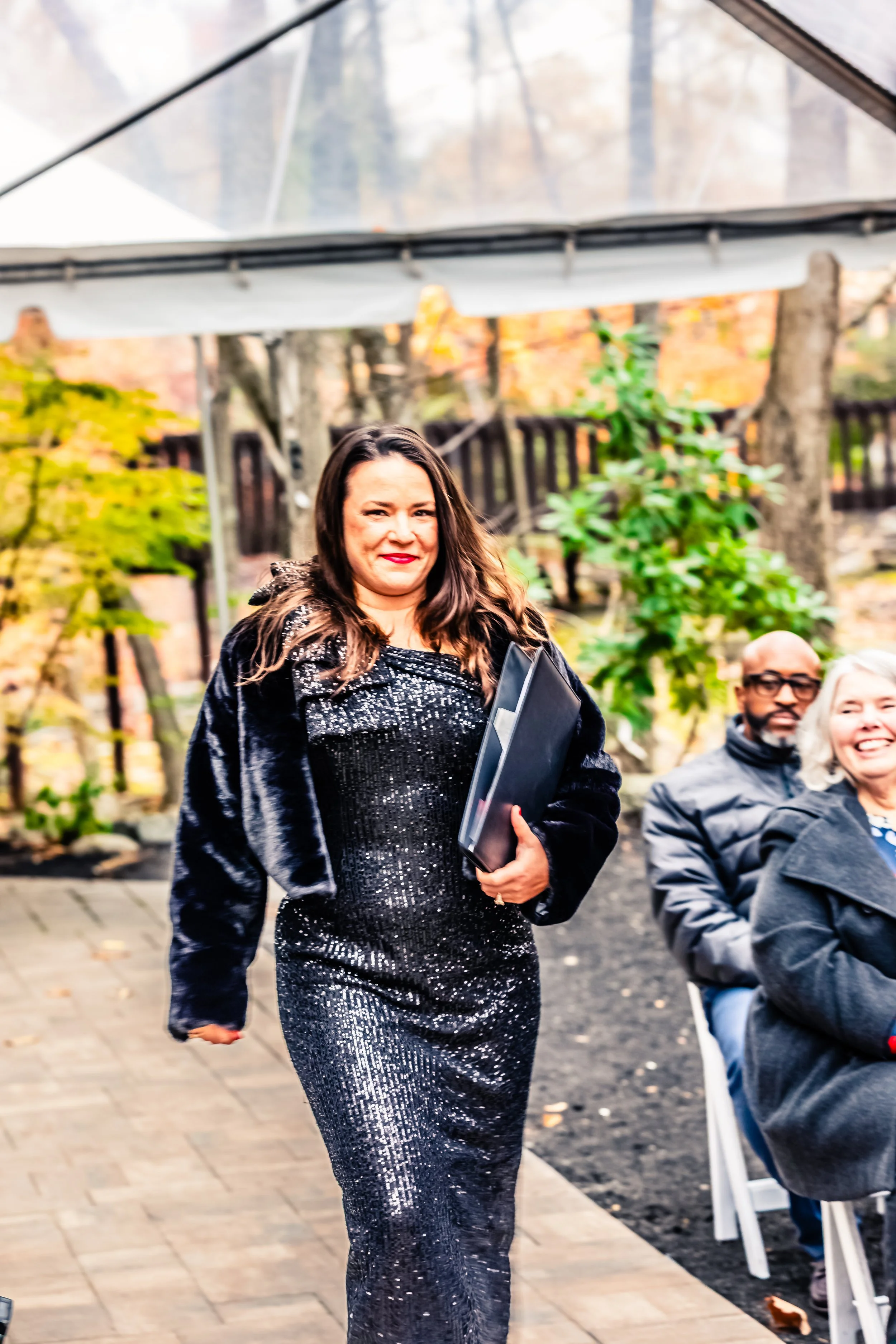 A woman wearing a black sequined dress and a black coat, holding a folder, walking outdoors with a smile. Two seated people and trees are visible in the background.