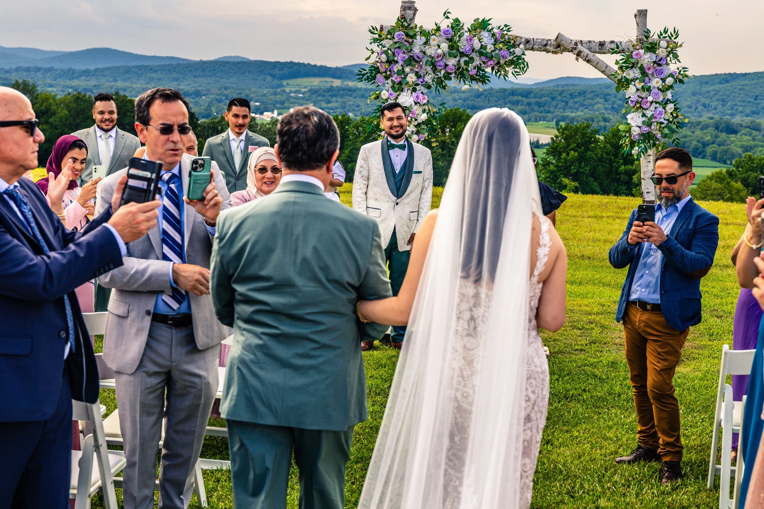 Outdoor wedding ceremony with a bride in a lace wedding gown and veil, and a groom in a white tuxedo with blue accents, standing beneath a floral arch, surrounded by guests taking photos and videos on their phones, with rolling green hills in the bac