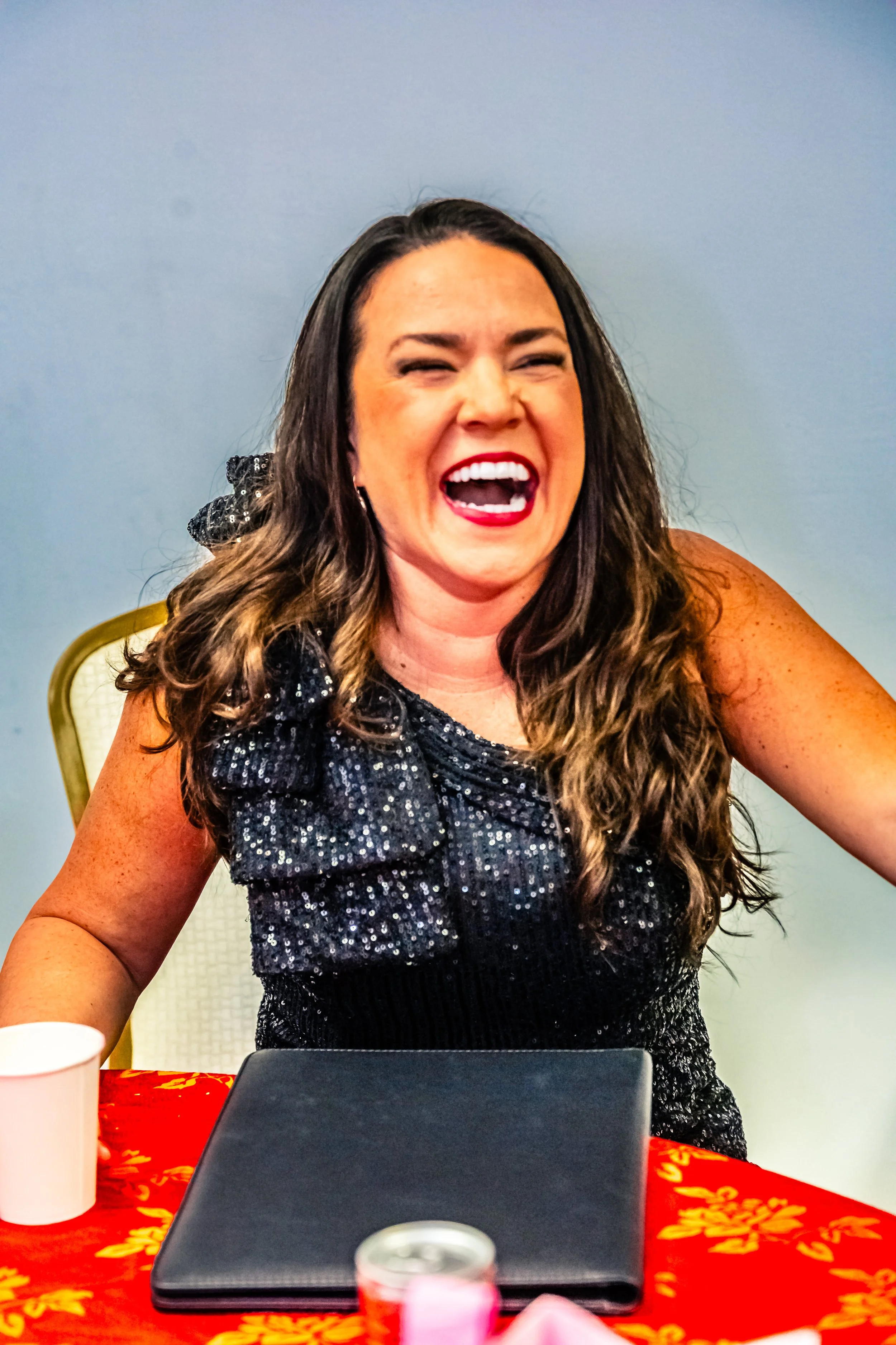 A woman with long, wavy dark hair, wearing a sparkly black dress, laughing with her mouth open, sitting at a table with a red and gold tablecloth, a closed laptop, a white cup, and a pink object in front of her.