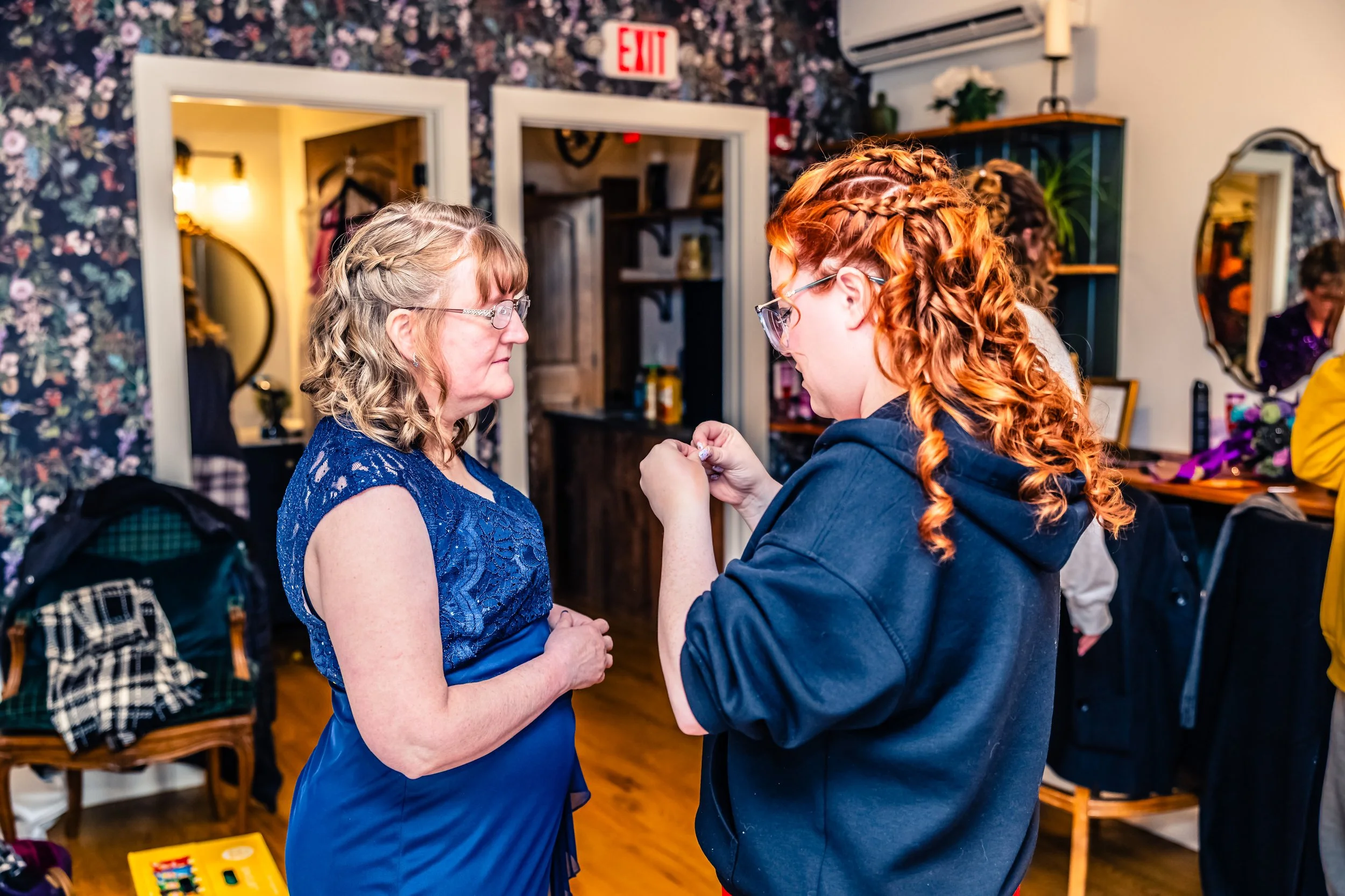 A woman with blonde hair, glasses, and a blue lace dress stands facing a younger woman with curly red hair, glasses, and a gray hoodie inside a room with floral wallpaper. The younger woman appears to be pinning or adjusting an item on the older woma