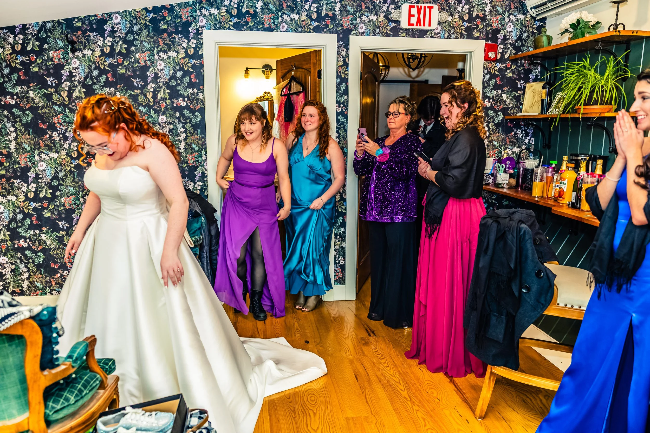 A bride in a white wedding gown stands on a wooden floor, surrounded by excited women dressed in colorful gowns. Some women are smiling and taking photos, while others watch the bride. The room has dark floral wallpaper, shelves with plants and bottl