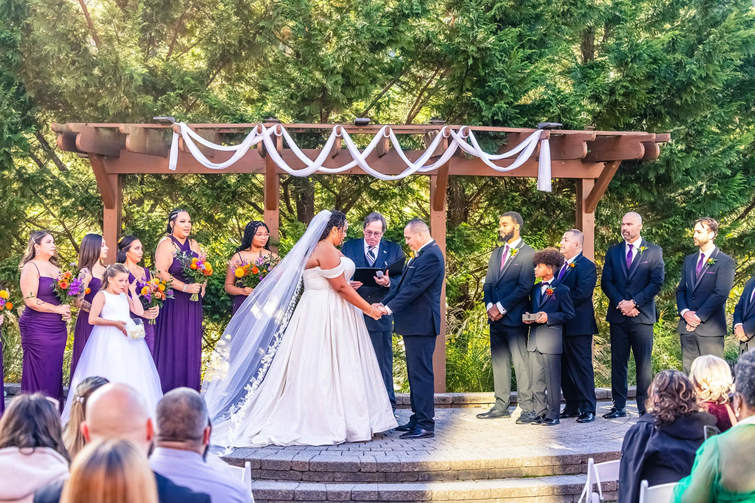 A wedding ceremony taking place outdoors with a bride and groom holding hands at the altar, surrounded by bridesmaids and groomsmen, with guests seated in front, under a wooden arch draped with white fabric, and lush green trees in the background.