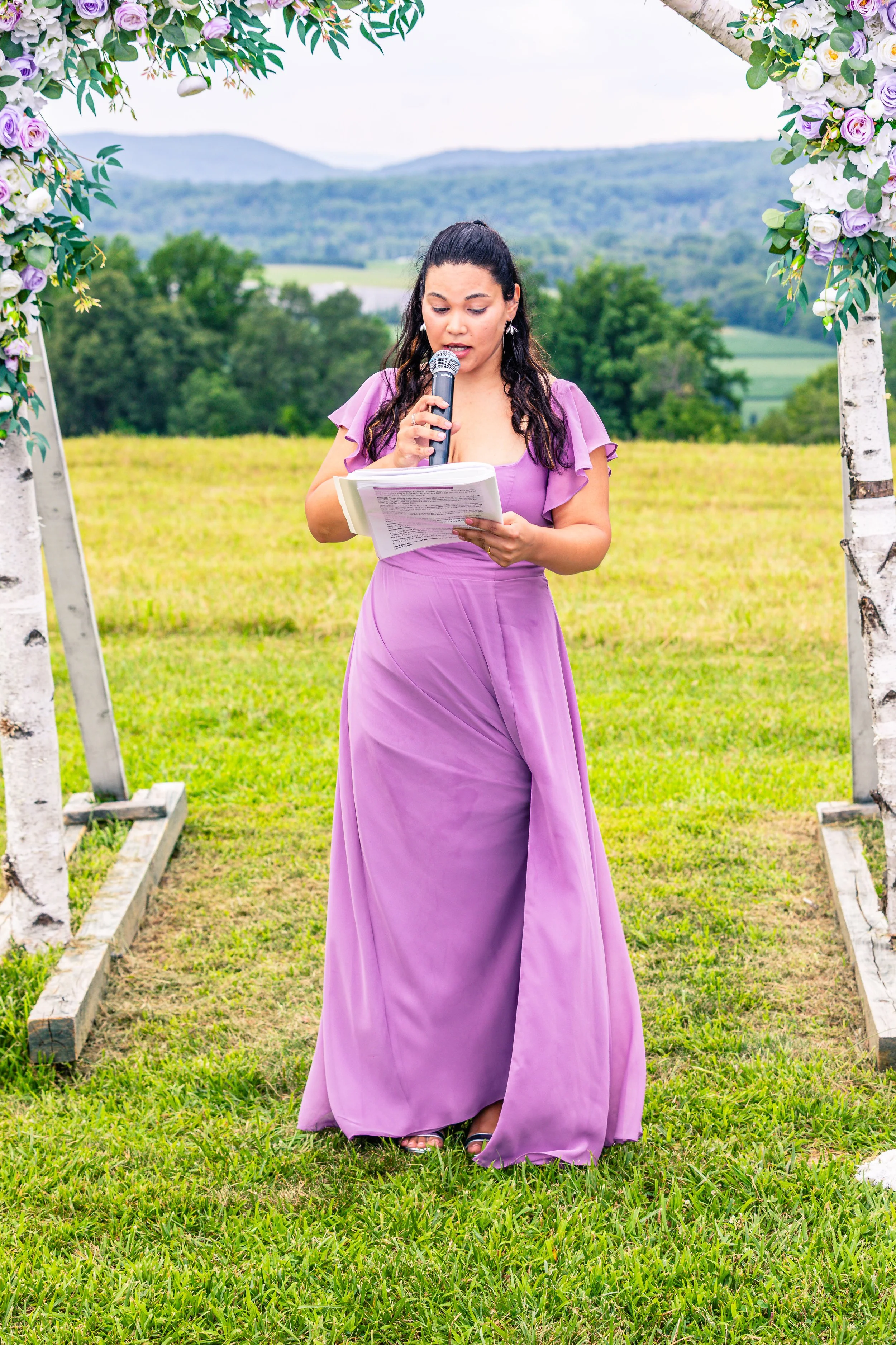 A woman in a purple dress stands outdoors, reading from papers into a microphone, surrounded by a floral arch at a scenic park with rolling hills in the background.