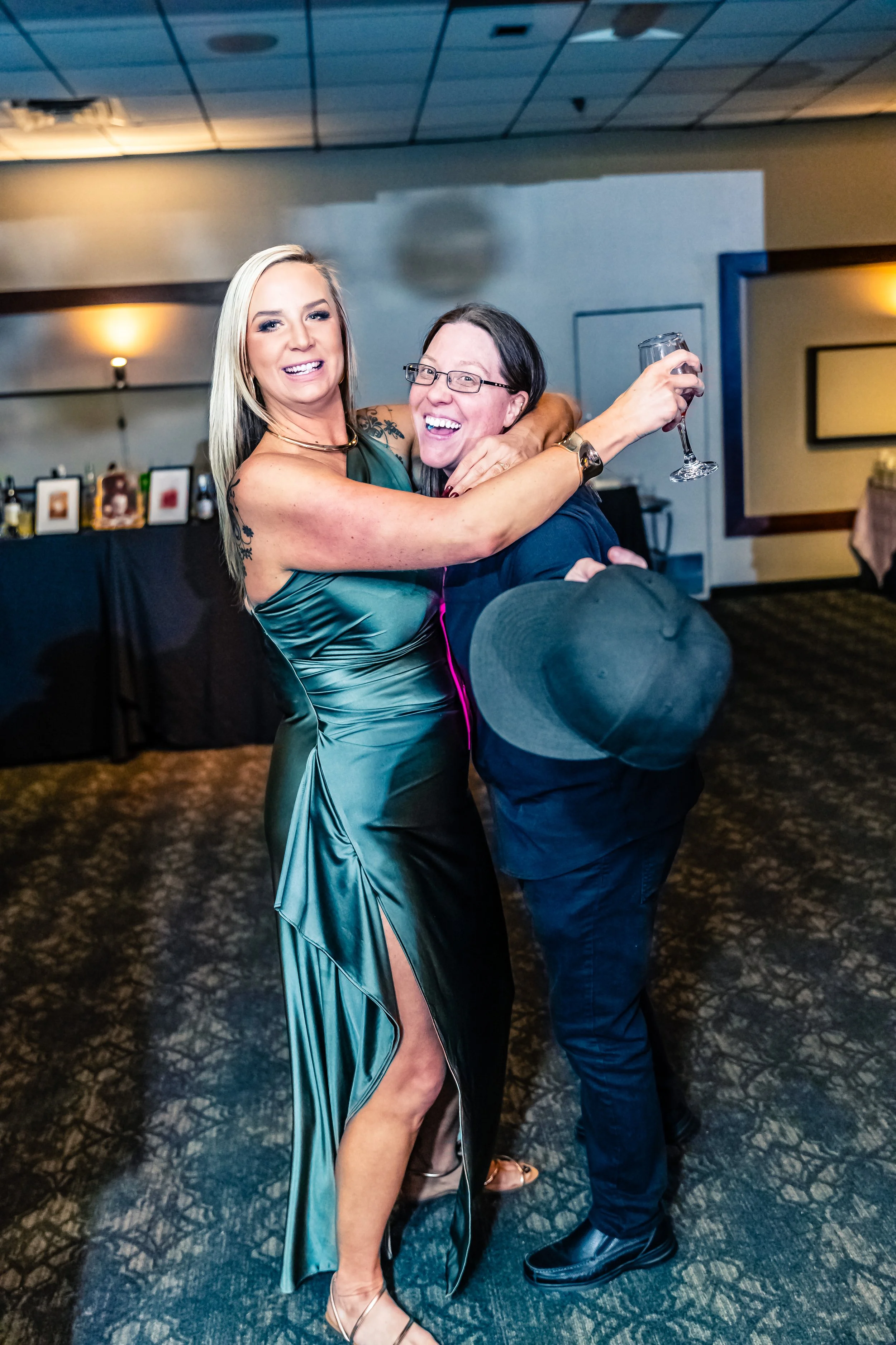 Two women hugging and laughing at a party, one holding a champagne glass, in an indoor event space.